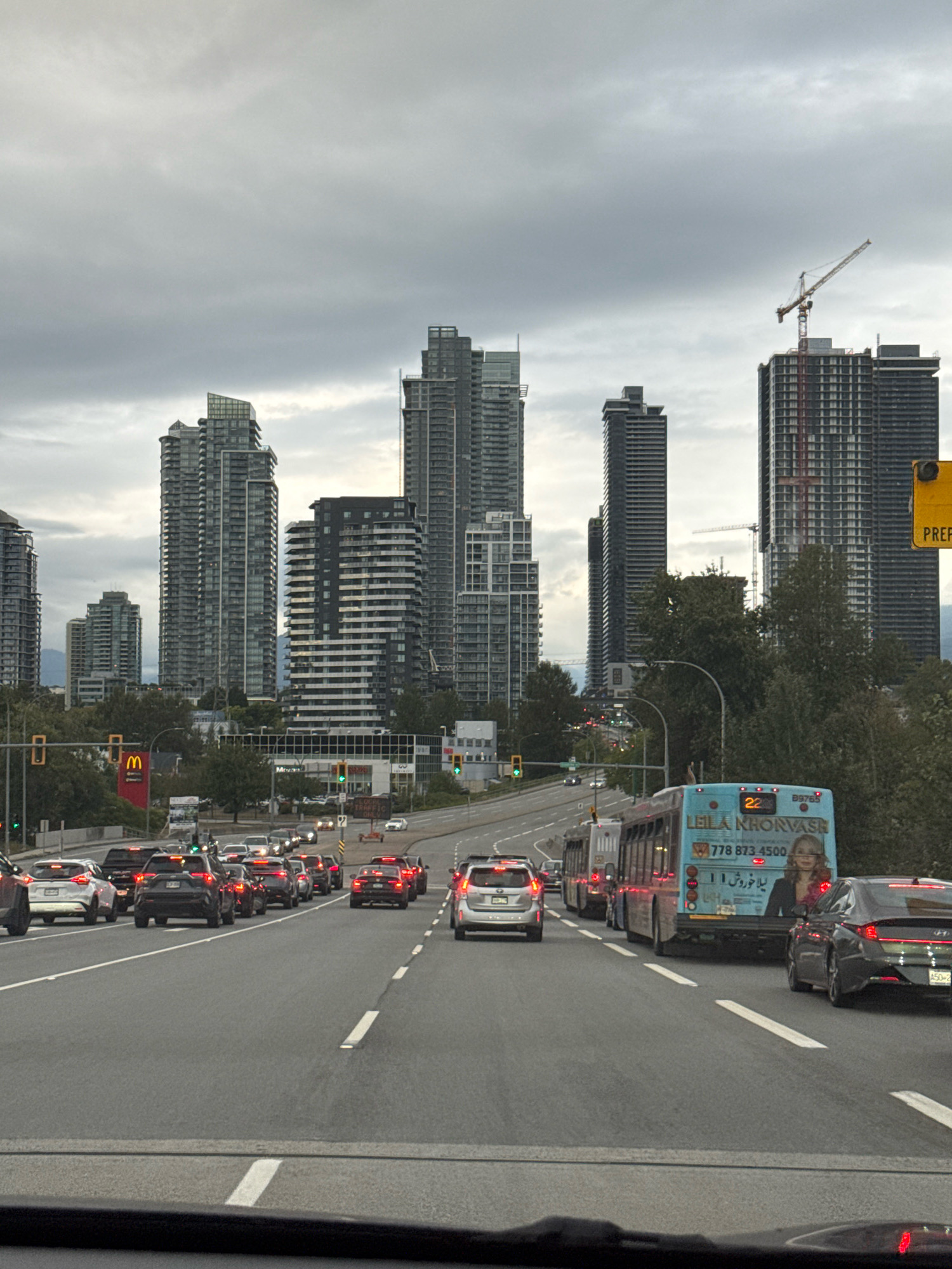 A wide shot captures a congested urban roadway in Burnaby, Canada, under an overcast sky. Numerous vehicles, including cars, SUVs, and buses, are stalled in traffic on a multi-lane highway. Several modern, high-rise buildings dominate the cityscape in the background, with a construction crane visible near one of the towers, indicating ongoing development. A bright yellow sign with black text is affixed to a pole on the right side of the image. On the left side, a red sign with a golden arches logo, characteristic of McDonald's, is prominently displayed. Traffic lights are visible, showing green signals. The foreground is dominated by the road surface and the bumpers of vehicles. The overall scene depicts a typical urban rush hour in a modern city.