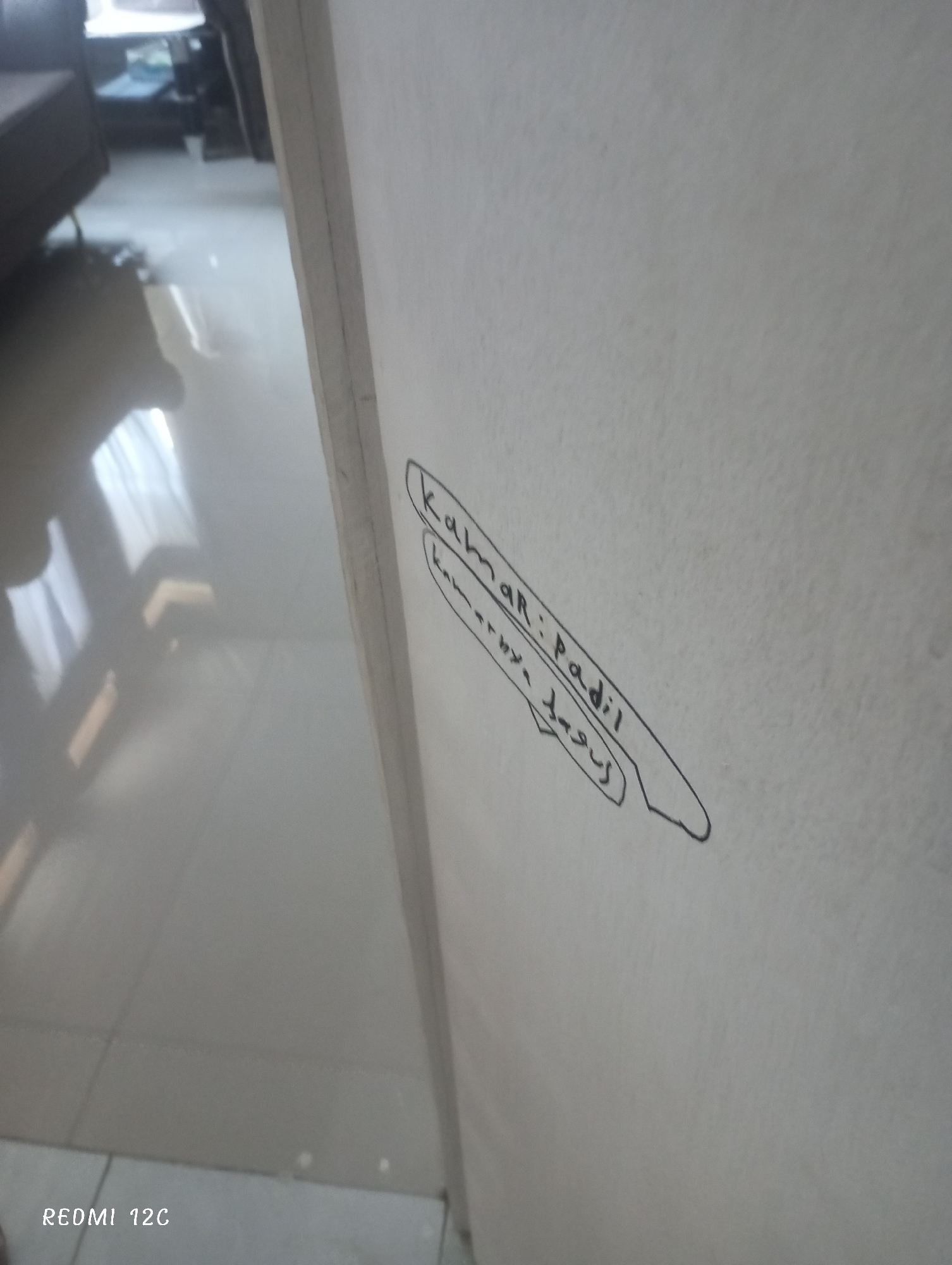 The image shows a close-up of a white wall with writing on it. The writing, surrounded by a thin, curved line, appears to say "Kumar: Padil" and "Kamarnya Bagus". The photograph was taken inside a building. No further information about the location in Lhokseumawe, Indonesia, can be determined from the image.