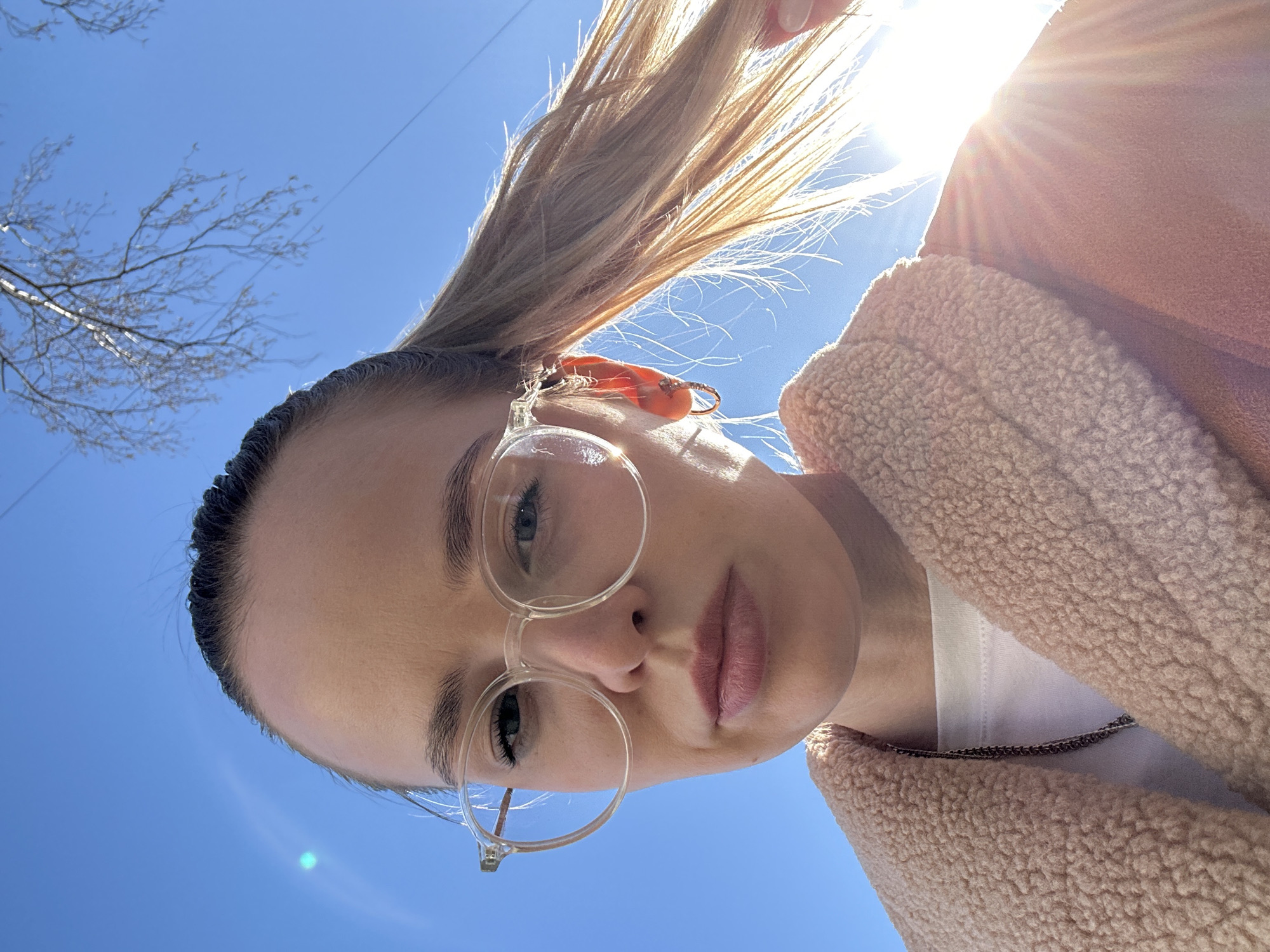 A low-angle, upward-facing photograph captures a woman with light skin and blonde hair pulled back in a high ponytail, wearing transparent-rimmed glasses. She is wearing a light pink, fuzzy jacket. The background features a clear blue sky with the sun positioned at the top, creating a lens flare effect. Bare tree branches are visible on the left side of the frame. The location is Chaikovsky, Russia.