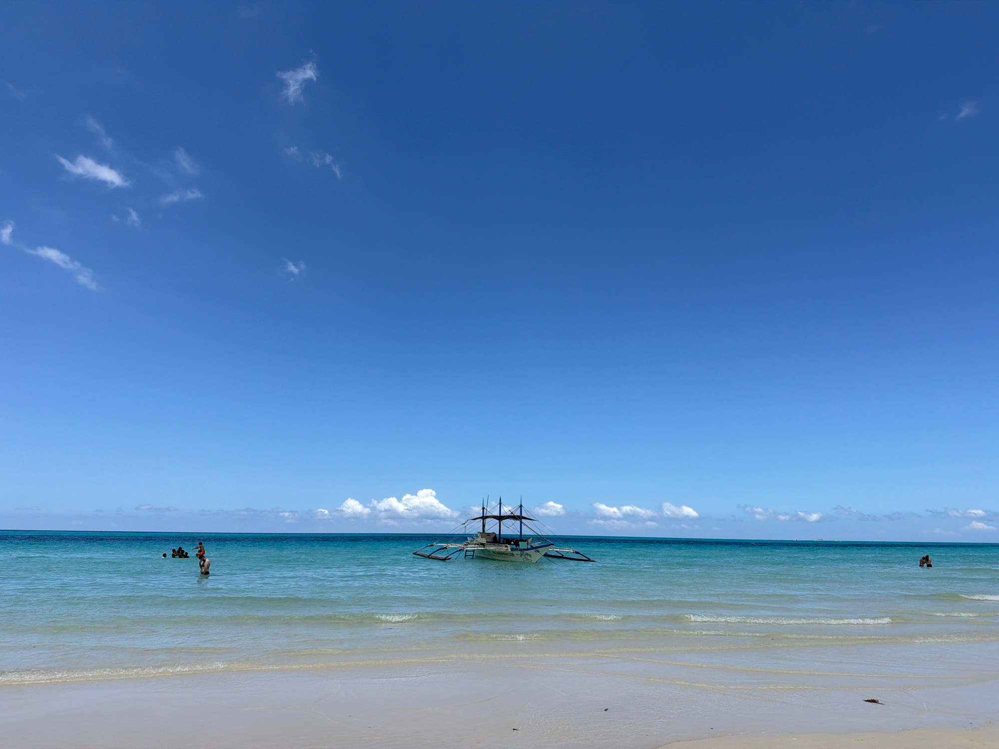 A bright, sunny day at a tropical beach in Malay, Philippines. The image captures a wide expanse of the sea and sky. The foreground shows a sandy beach with gentle waves lapping at the shore. The water is a clear, turquoise blue, transitioning to a deeper blue towards the horizon.
In the middle ground of the water, a traditional Filipino outrigger boat (also known as a 'paraw') is anchored. The boat has multiple masts and outriggers, suggesting it is ready for sailing or is a leisure craft. A few scattered clouds dot the clear blue sky, casting soft shadows on the water.
On the left side of the image, in the shallow water, a group of people are visible. Some are standing, appearing to be swimming or enjoying the water. One individual is wearing an orange shirt. Further to the right, a single person is visible in the water, and to the far right, two people are in the sea. The overall scene depicts a serene and inviting beach environment.