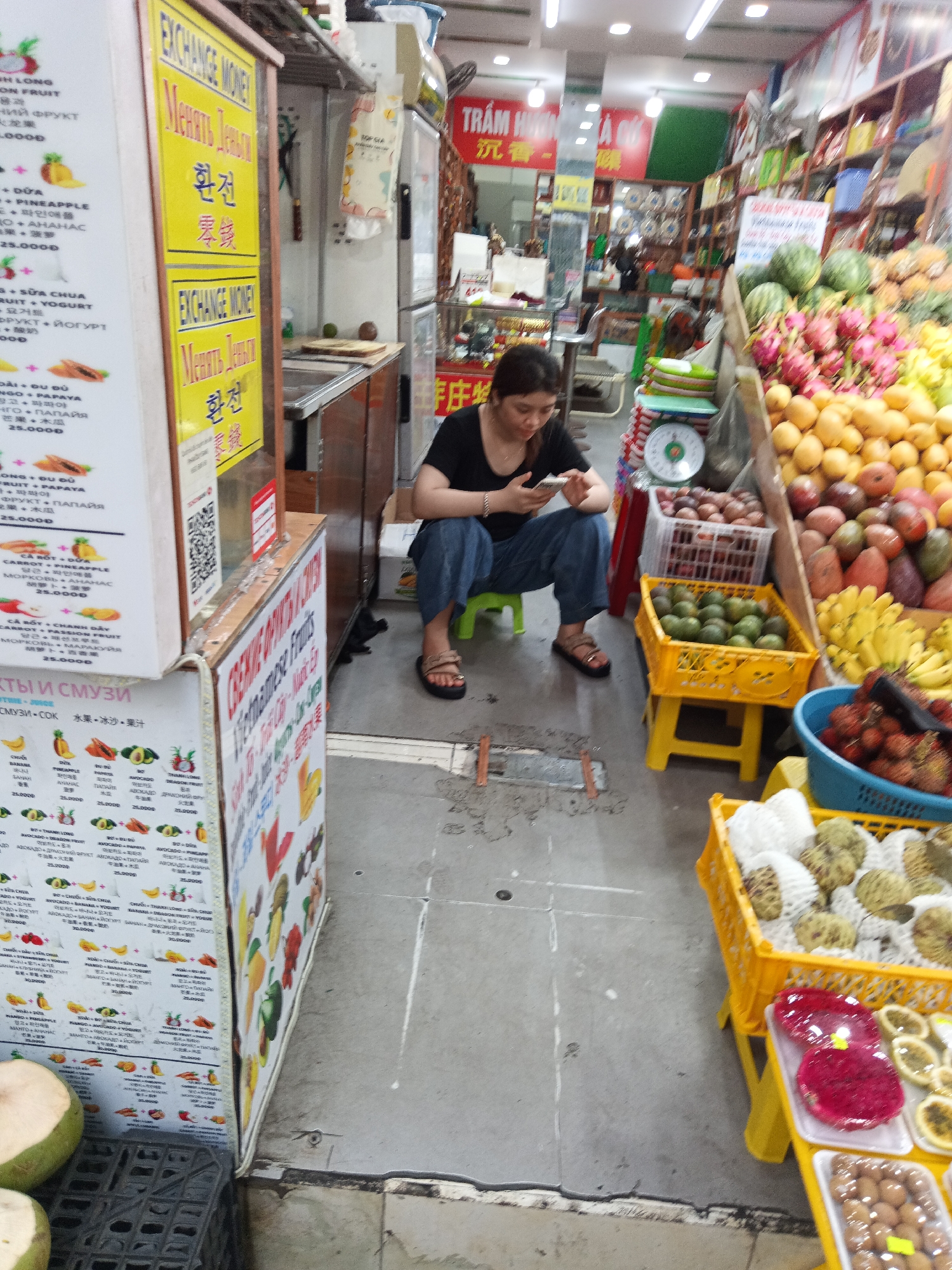 The image shows a woman sitting on a small green stool in a busy market in Nha Trang Ward, Vietnam. She is engrossed in her smartphone. Stalls overflowing with various fruits, including mangoes, dragon fruit, avocados, and bananas, are densely packed around her. Signs displaying fruit smoothie menus and exchange rates in multiple languages adorn the stalls. The market atmosphere is bustling, with people and goods crammed into a relatively small space.