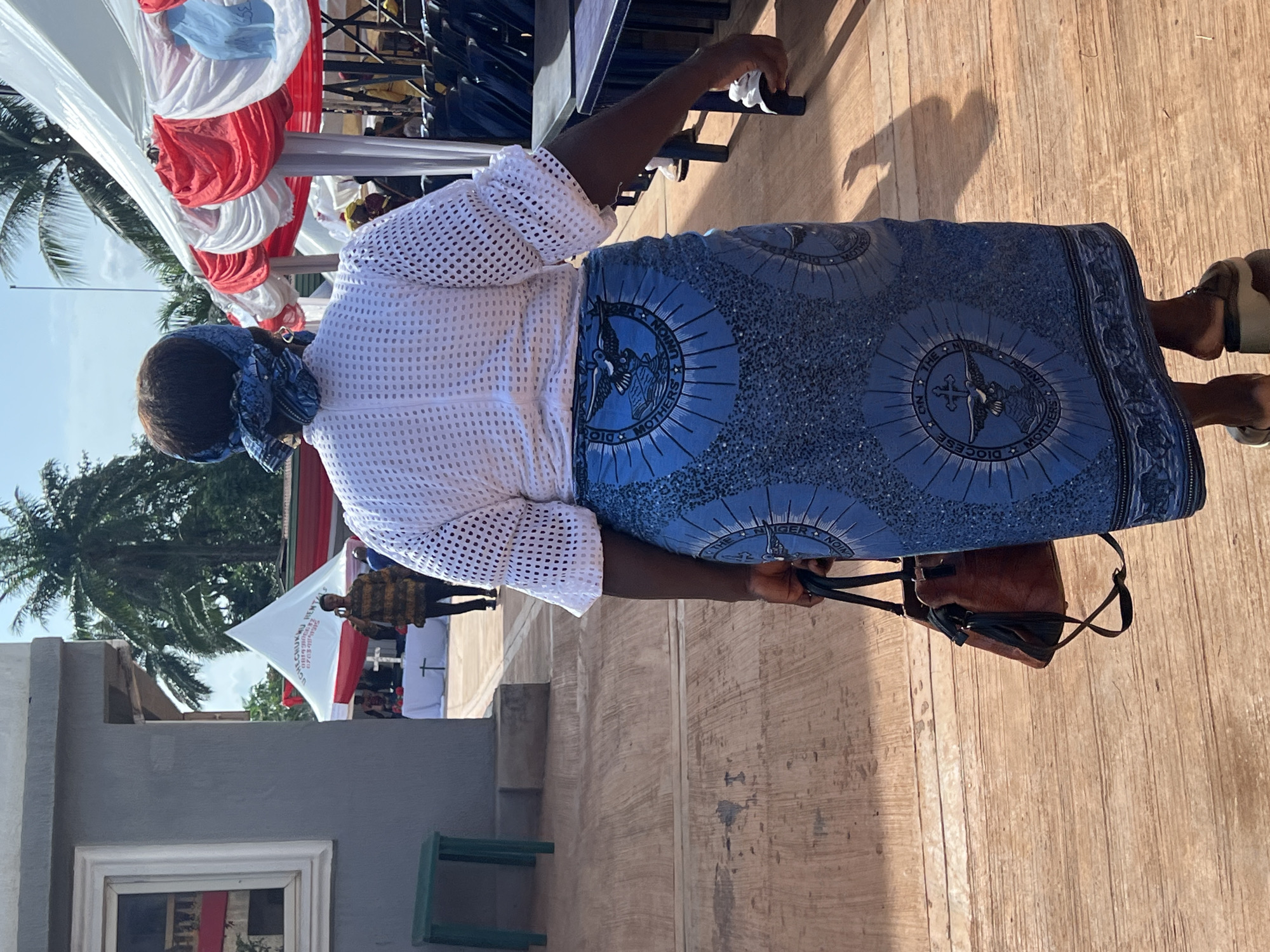 The image captures an outdoor event in Onitsha, Nigeria, on a bright, sunny day. The primary subject is a person, likely a woman, seen from behind and to the side, standing on a light brown, possibly wooden, paved surface. She wears traditional attire: a white, open-knit long-sleeved top and a vibrant blue long skirt. The skirt is decorated with circular patterns, each featuring a logo that includes a cross, a dove-like figure, and the text "DIOCESE MOTHERS' UNION". Other partial text on the skirt reads "THE MOTHERS' UNION" and "NIGER NOIN," likely referring to "Nigeria Union." Her head is covered with a blue patterned fabric, and she carries a brown handbag in her left hand while wearing light-colored sandals.
In the background, there's an event setup with a white canopy draped with red and white fabric. Palm trees and a clear sky indicate a tropical or warm climate. A white banner or flag is visible, bearing text such as "UCHECHUKKI," "08199100," "HENTAS," "COMMUNION RENTAL," and "THE MINDER." Distant figures and structures, including a green stool, suggest a public gathering. The attire and the prominent "Mothers' Union" insignia on the skirt point to participation in a religious or community-based organization's event.