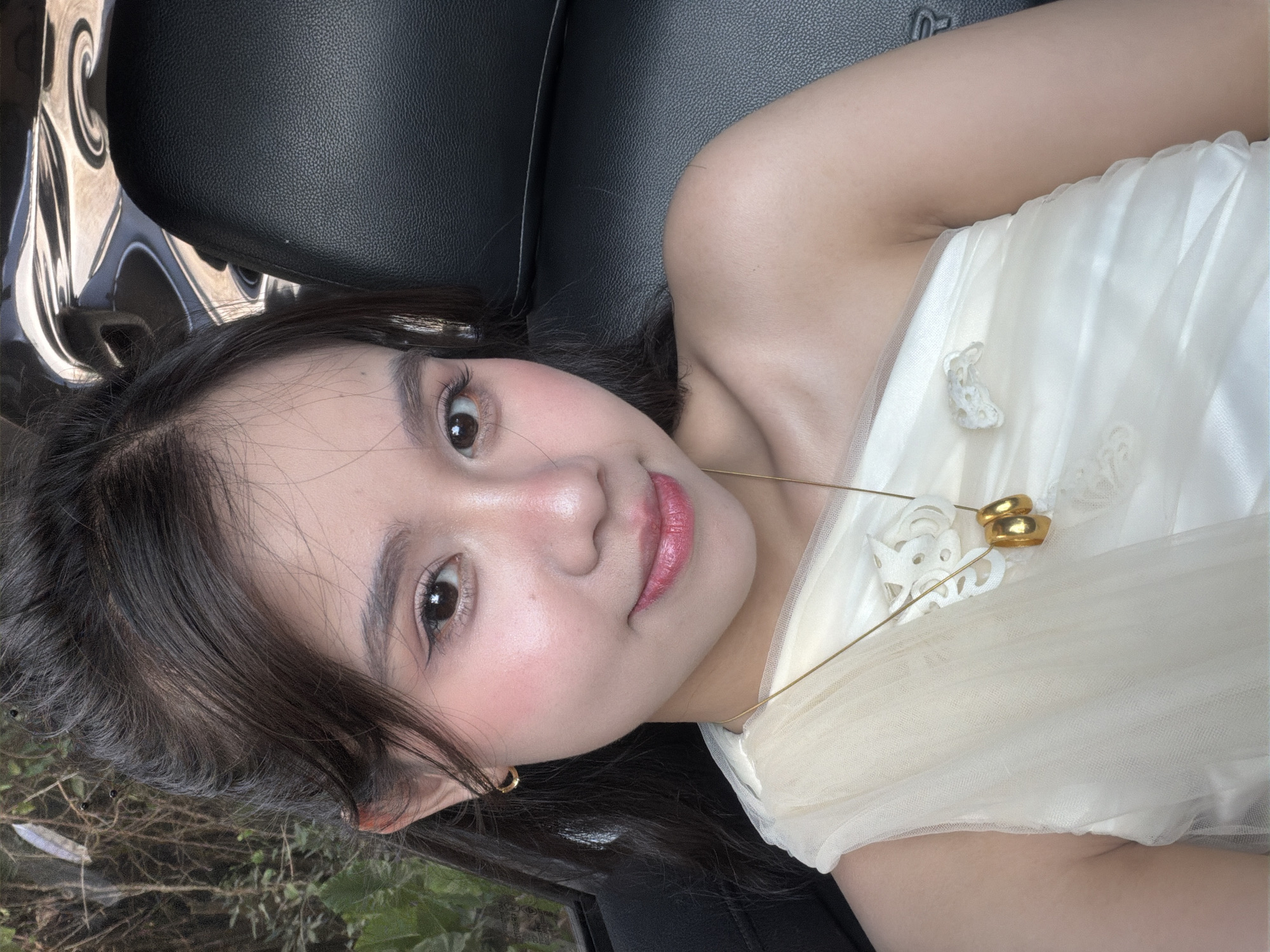 A young woman with long, dark hair and fair skin is lying down, possibly in a car. She is wearing a white dress with a delicate, lacy design and a gold necklace. The background appears to be a dark car interior with a glimpse of outside scenery, potentially foliage. No specific details indicate the city of Bình An, Vietnam, so that aspect cannot be confirmed.