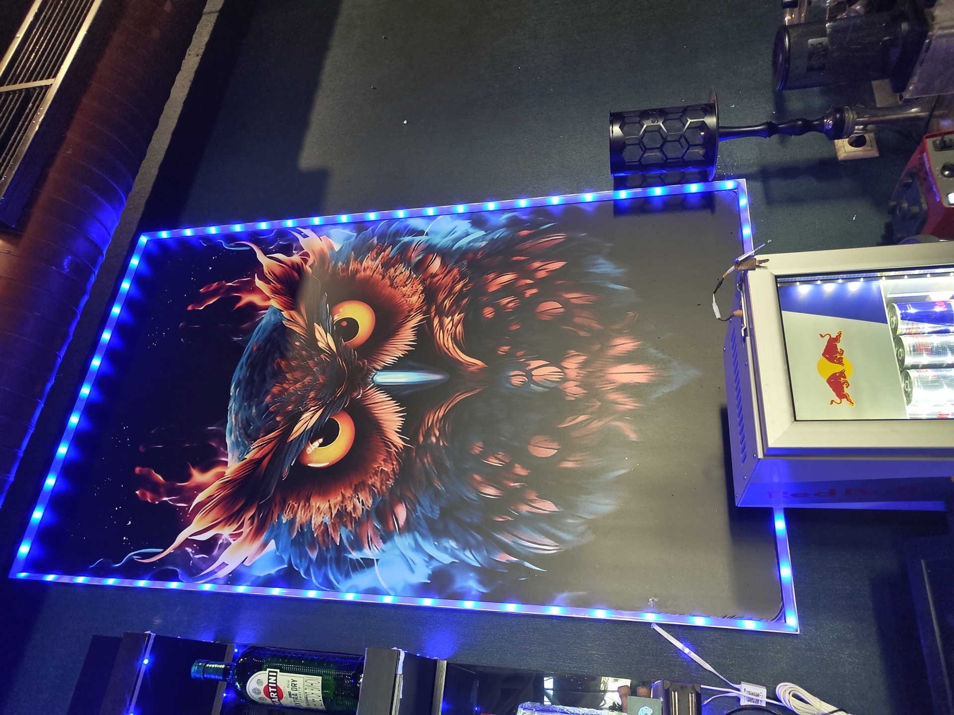 In an indoor setting, a large, striking poster of an owl with fiery plumage is displayed on a dark wall, illuminated by blue LED strip lights outlining its border. To the left of the poster, a green bottle of Martini Extra Dry vermouth stands on a shelf. Below the poster, a compact, white mini-fridge branded with the Red Bull logo is visible, containing cans of what appears to be Red Bull. To the right of the fridge and poster, a black hookah with a decorative stem stands next to a small, black speaker with a honeycomb pattern. The scene appears to be a bar or lounge area. The city of Zhukovsky, Russia, is not visually represented in this image.