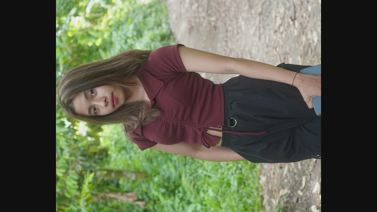 A young woman with long, brown hair stands on a dirt path in a wooded area. She is wearing a maroon, ruched crop top with a collar and short sleeves, and black cargo pants. The woman is holding a light blue cellphone in her right hand. The background consists of green foliage and trees.