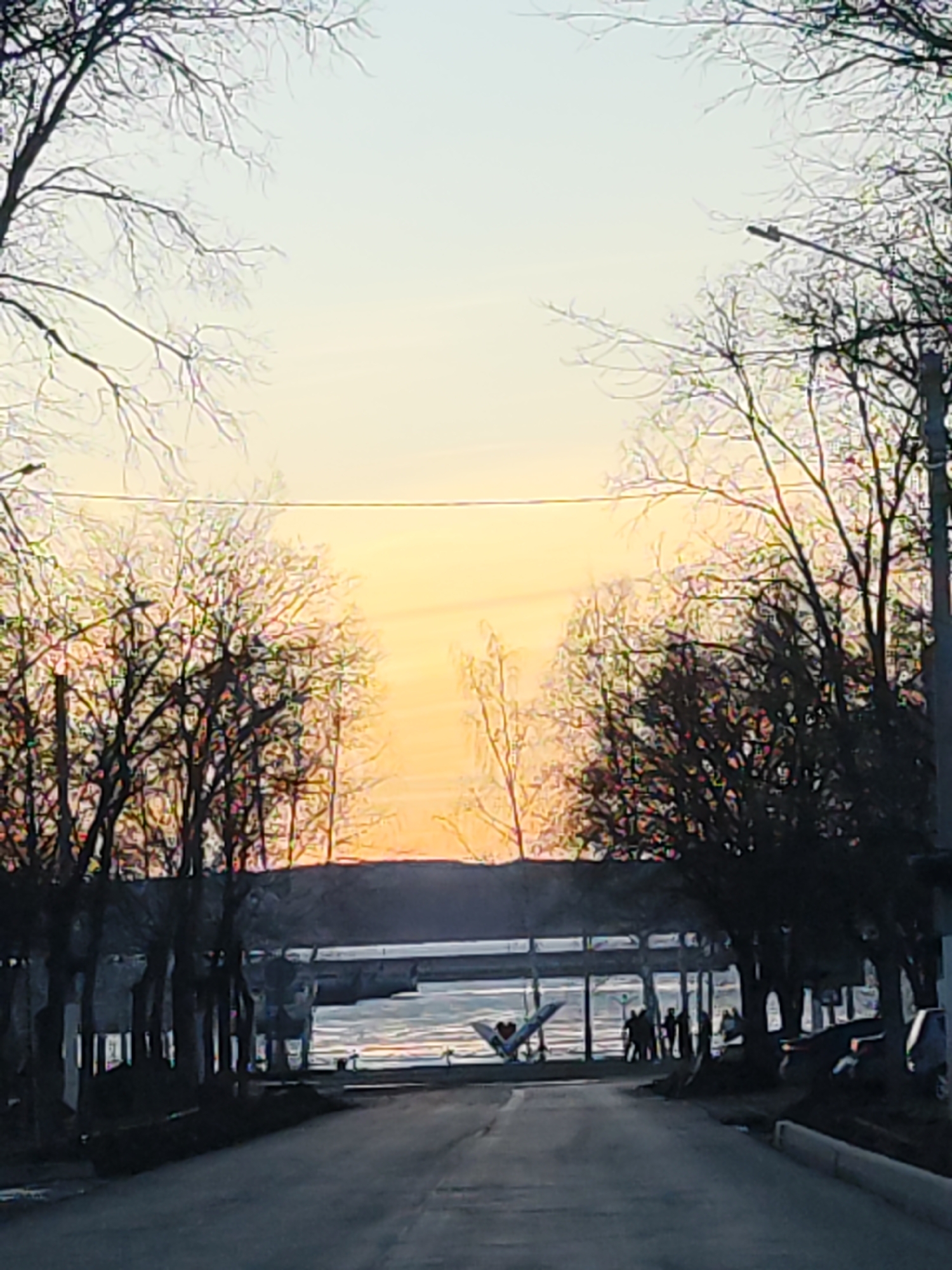 A street scene in an urban location, possibly near water, is depicted at dusk or dawn. The road leads toward a waterfront area where people are gathered. Bare trees line the sides of the street, indicating a colder season. A warm, orange glow suggests a sunrise or sunset, and the light is reflected on the water. A couple of cars are parked to the right of the road. A structure, possibly a building or pavilion, is partially visible behind the gathering of people near the water.