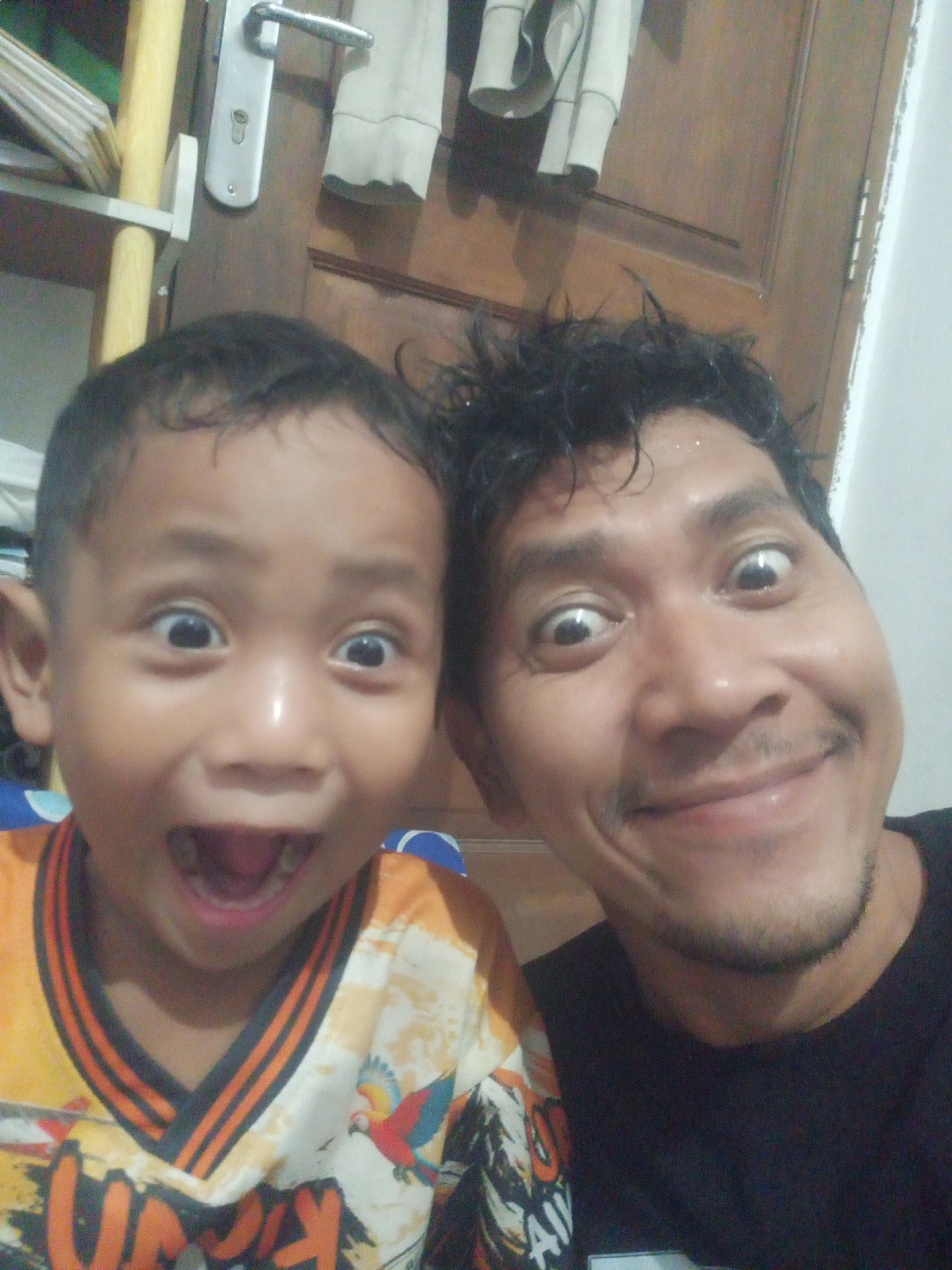 A selfie taken indoors shows two males, an adult and a child, with their faces close to the camera. The adult, on the right, has dark, disheveled hair and a wide-eyed expression. He is wearing a black t-shirt. The child, on the left, has short, dark hair and is also making a surprised facial expression, with a wide-open mouth. He is wearing an orange and black collared shirt with a parrot design. In the background, a wooden door with a silver handle is visible, along with a shelf holding some items. The lighting suggests an indoor setting, likely a home. There are no identifying markers to place this image specifically within East Jakarta.