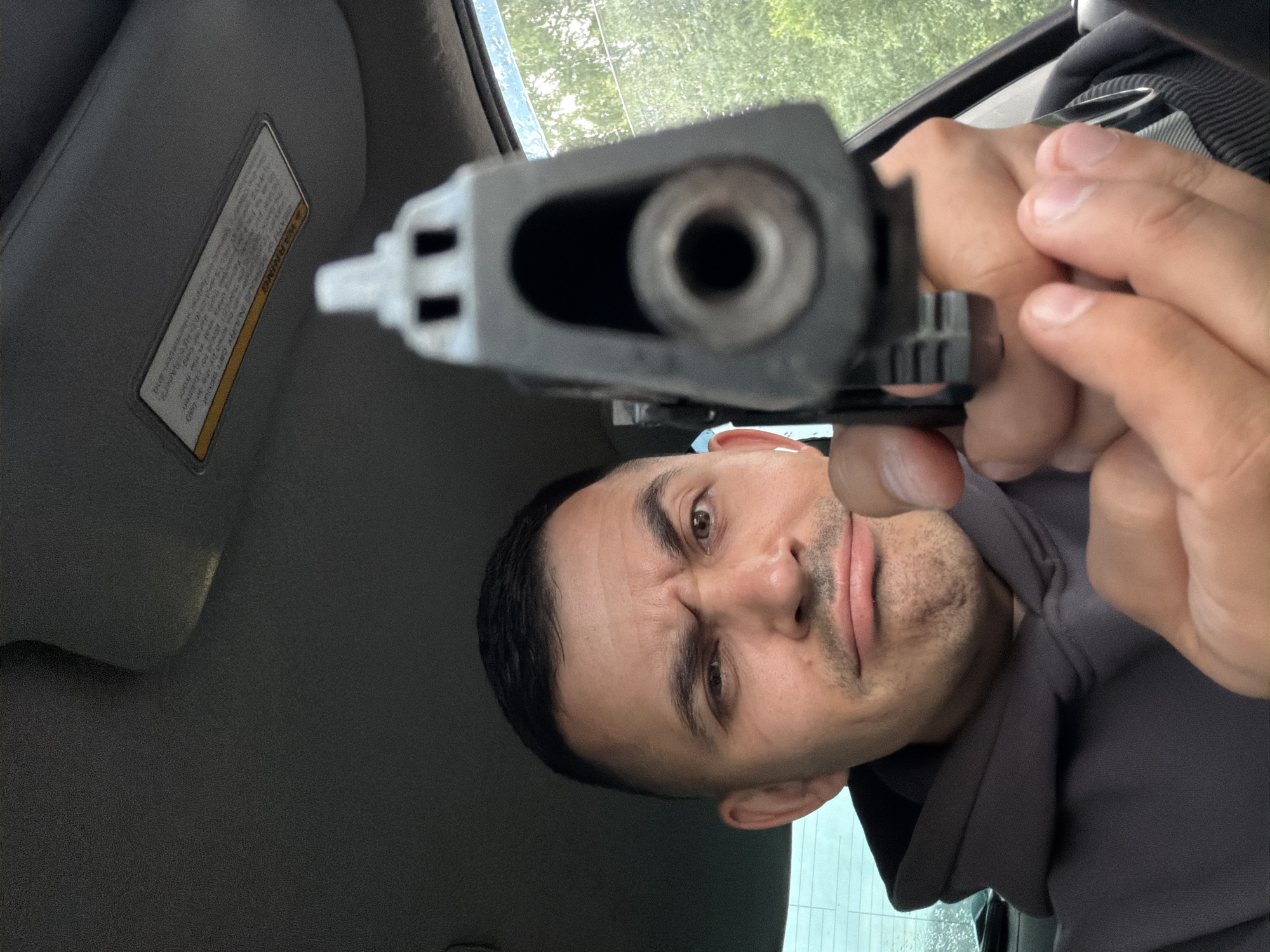 The image shows a person in a car, holding a handgun and pointing it towards the camera. The person appears to be male with short dark hair and a goatee. He has a stern expression on his face. The handgun is a dark color and is in focus, while the person's face is slightly blurred. The interior of the car is visible, including a sun visor with a warning label. The background shows greenery through the car window, suggesting an outdoor setting. The lighting appears to be natural, indicating daytime. There is no clear indication of the specific time of day or weather conditions beyond the greenery. The warning label on the sun visor has text, but it is not legible. The overall impression is one of confrontation or threat due to the pointing handgun and the person's expression.