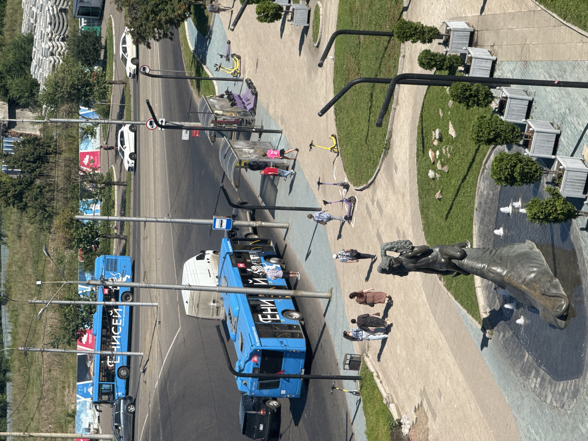 This is an aerial shot of a street scene in Krasnoyarsk, Russia, on a sunny day. The image features a blue bus, white van, and a black car on a paved road. To the right of the road, a wide pedestrian walkway is lined with grassy areas, trees in planters, and a fountain with a statue. Several people are walking on the pedestrian path, some near a bus stop with multiple scooters parked. The time of day appears to be midday, given the bright sunlight and strong shadows.
Text visible on the bus includes "ЧИСТЫЙ ДВИЖЕНИ" and what appears to be a logo with a zebra. There are also traffic signs and advertisements visible in the scene. The overall mood is one of urban activity on a pleasant day.