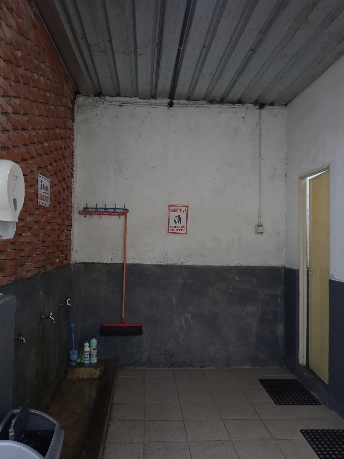 The image shows a small, enclosed space with a corrugated metal ceiling. The walls are painted white on the top half and dark grey on the bottom half. There is a small, white, rectangular sign with red lettering that says "PERHATIAN" (Indonesian for "Attention") and a small, black pictogram of a person walking. There is also a red, plastic broom leaning against the wall, a rack with a string of hooks above it, a white, plastic, wall-mounted dispenser, and a door with a white frame that is closed. The floor is tiled and there are a few pieces of dirt and debris scattered throughout. Two faucets are attached to the wall near the floor, and there is a drain running down the wall in the corner of the room. There is a gray trash can partially visible in the lower left-hand corner of the image. The space appears to be an outdoor bathroom or a utilitarian area used for cleaning. The coordinates -7.669, 111.217 indicate a location in the city of Plaosan, Indonesia.