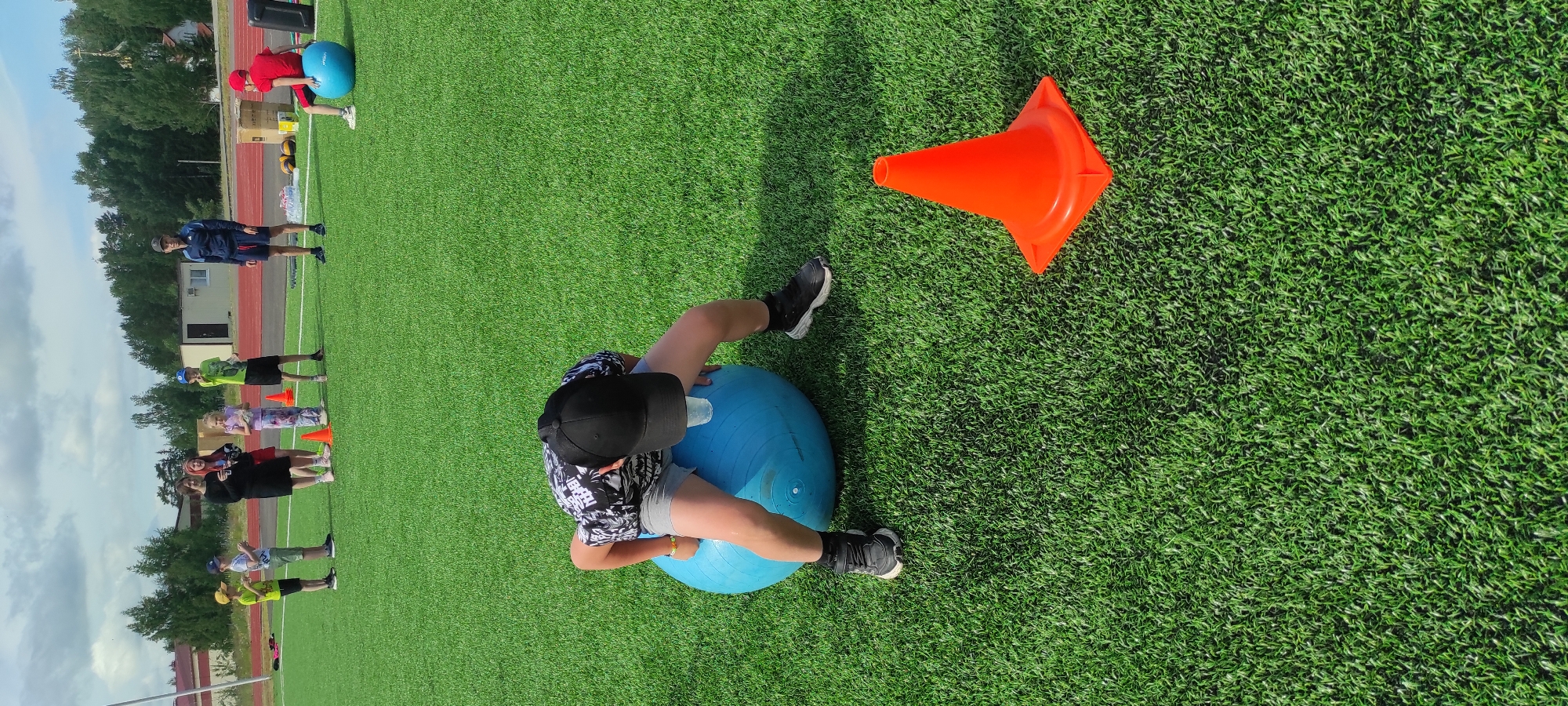 In Noviy, Russia, on a bright, overcast day, a group of people, predominantly children, are gathered on an artificial turf sports field. In the foreground, a young boy wearing a black baseball cap, a black and white patterned t-shirt, and grey shorts is crouched over a large, blue exercise ball. He appears to be balancing a clear plastic cup on his head while seated on the ball. His legs are bent, and his feet are planted on the grass on either side of the ball, with black athletic shoes. To the right of the boy and the exercise ball, an orange traffic cone stands on the turf. In the background, a few adults and other children are visible. One adult man in a blue tracksuit and shorts stands observing. Another man in a red shirt is seated on a smaller blue exercise ball to the far right. The field is lined with red track material and surrounded by trees and buildings under a cloudy sky.