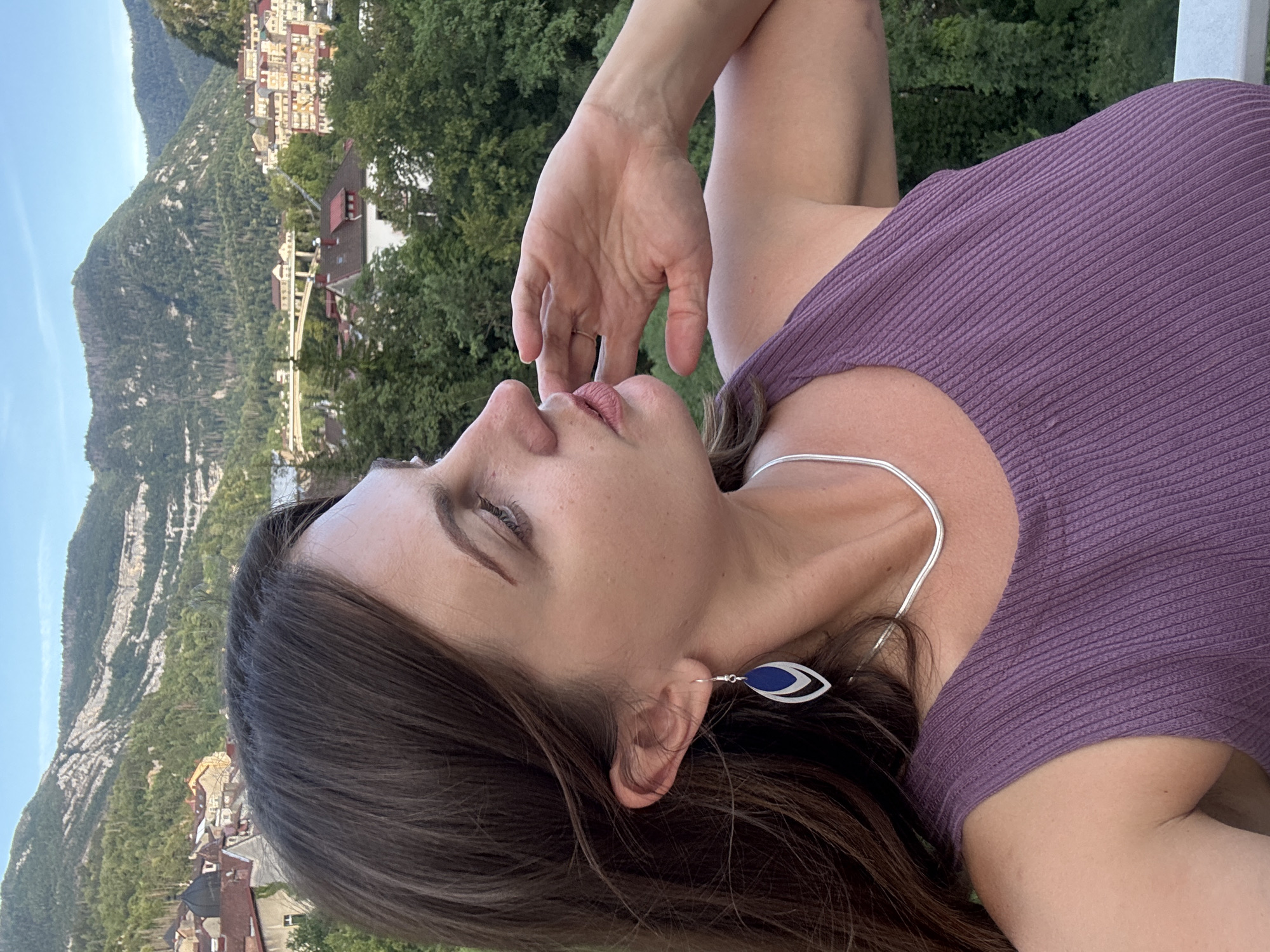 The image captures a woman with long, dark brown hair, wearing a purple tank top, a silver snake chain necklace, and a teardrop-shaped earring with silver and blue accents. She is looking to her right, with her right hand touching her face near her mouth. Her skin tone is fair, and she has light makeup on.
The background reveals a scenic view of Saint-Claude, France. The town is nestled in a valley, with buildings featuring red-tiled roofs and cream-colored facades. A bridge spans across a distant part of the town, and lush green trees dominate the mid-ground. In the background, steep, tree-covered mountains rise under a clear blue sky. The lighting suggests it is late afternoon or early evening.
