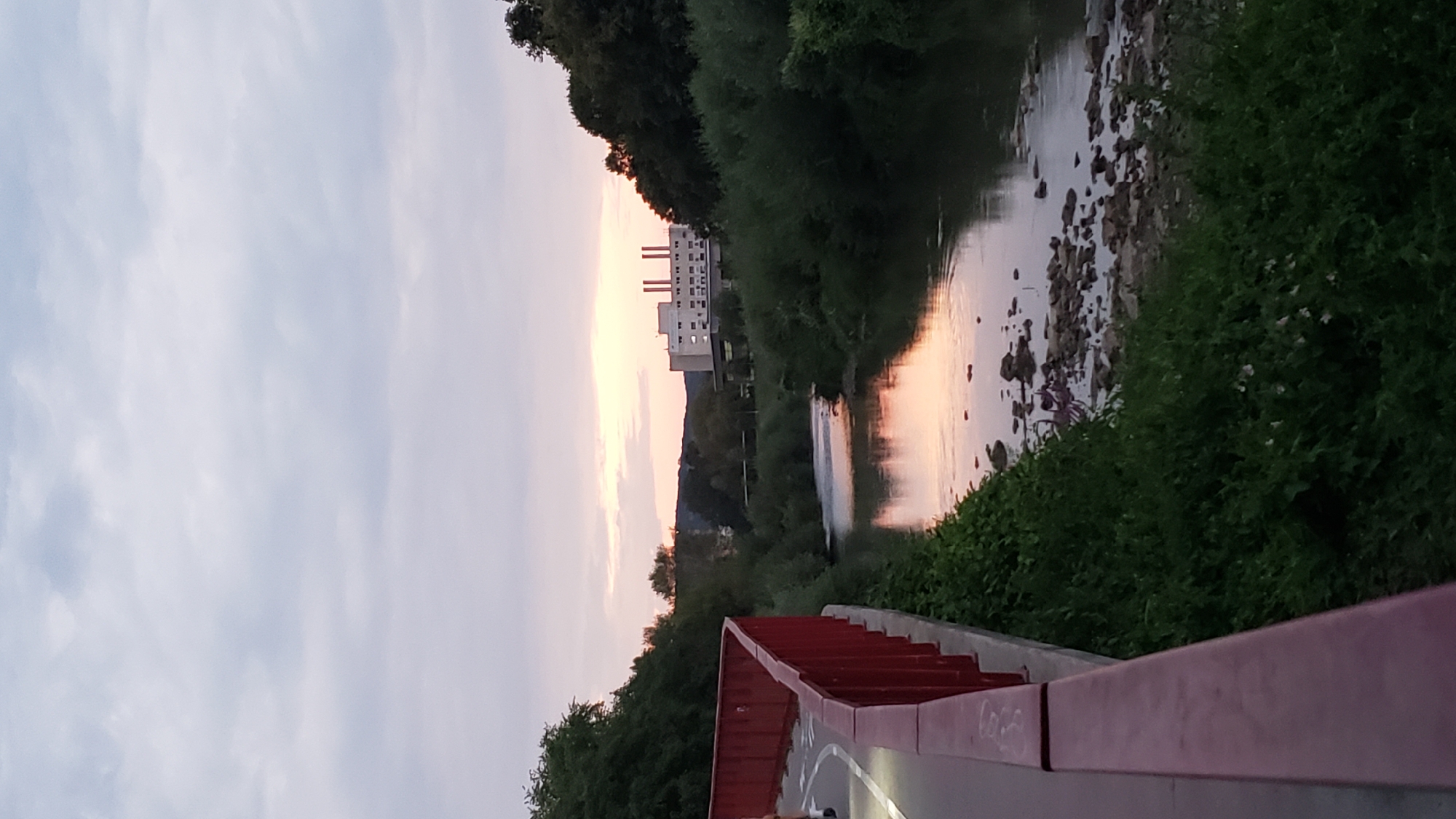 A vertical, eye-level shot captures a tranquil sunset scene along a riverbank in Prešov, Slovakia. On the left, a bright red railing lines a paved walkway, with a portion of a person's legs visible, suggesting they are walking or running. The walkway curves out of frame to the left. To the right of the walkway, lush green vegetation grows densely, sloping down towards a calm river. The river's surface reflects the soft hues of the twilight sky, with patches of pink and orange indicating the setting sun, which is just visible peeking through the clouds near the horizon. In the background, across the river, is a large, light-colored building with multiple windows and twin smokestacks, characteristic of an industrial or institutional facility. The building is surrounded by trees and foliage, and a gently rolling, tree-covered hill forms the distant backdrop under a pale, cloudy sky. The riverbed is visible in places, with a scattering of rocks.