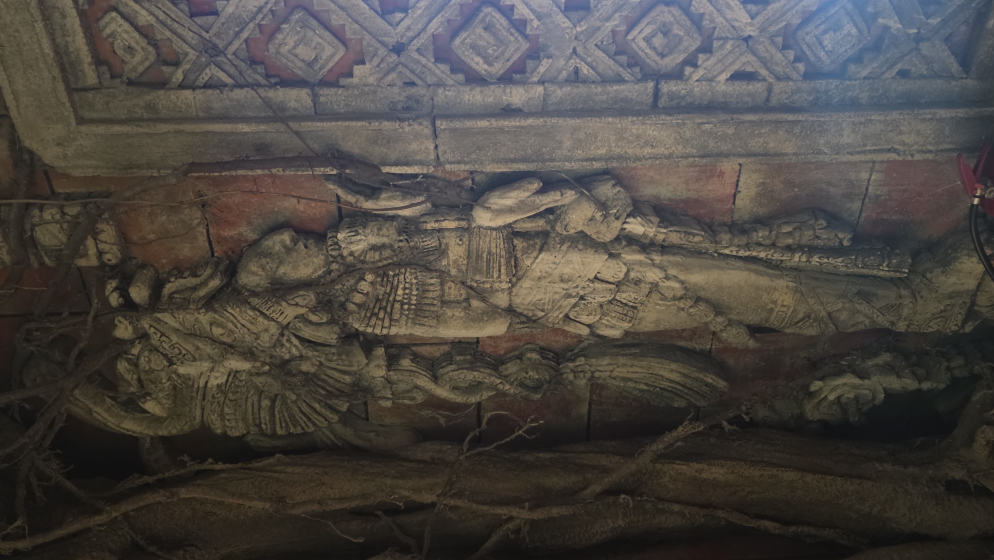 The image depicts a stone carving of a reclining figure, adorned with detailed decorative elements, positioned beneath a patterned ceiling. The environment appears weathered and possibly ancient, with roots or vines adding to the sense of age. Based on its style, the carving is reminiscent of Mayan or Aztec artwork, possibly from a temple or ceremonial site. The precise location within an unknown city is indeterminable without more context.