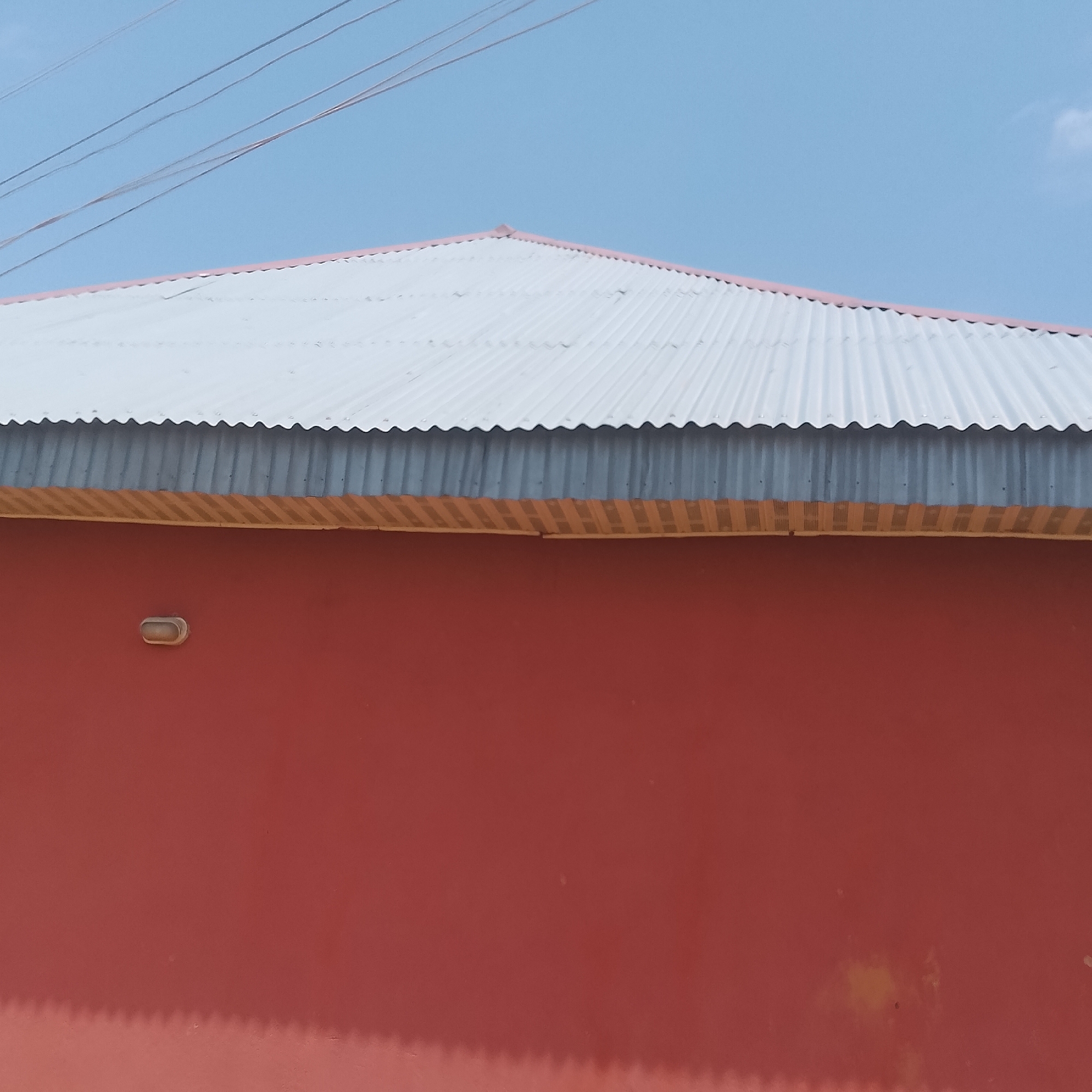 An outdoor, eye-level, medium shot shows the exterior of a building under a clear blue sky with a few wispy clouds. The top of the image features a corrugated metal roof that is light gray in color and slants downwards from left to right. Below the roofline, a band of metal siding runs horizontally across the width of the image. The siding is a muted gray color and exhibits a ribbed texture. The lower portion of the image is dominated by a solid red wall, likely painted stucco or concrete, which appears to be the main facade of the building. A small, oval-shaped light fixture is mounted on the red wall, slightly off-center to the left. The lighting suggests it is daytime, and the shadow cast by the roof overhang on the red wall is visible, indicating a shadow line with a serrated edge, mirroring the waves of the corrugated metal. There are several thin, dark lines crisscrossing the sky in the upper left corner of the frame, possibly power lines or wires. The overall scene depicts a simple building structure in an urban or semi-urban environment, possibly in Jalingo, Nigeria, given the context. There are no people visible in the image.