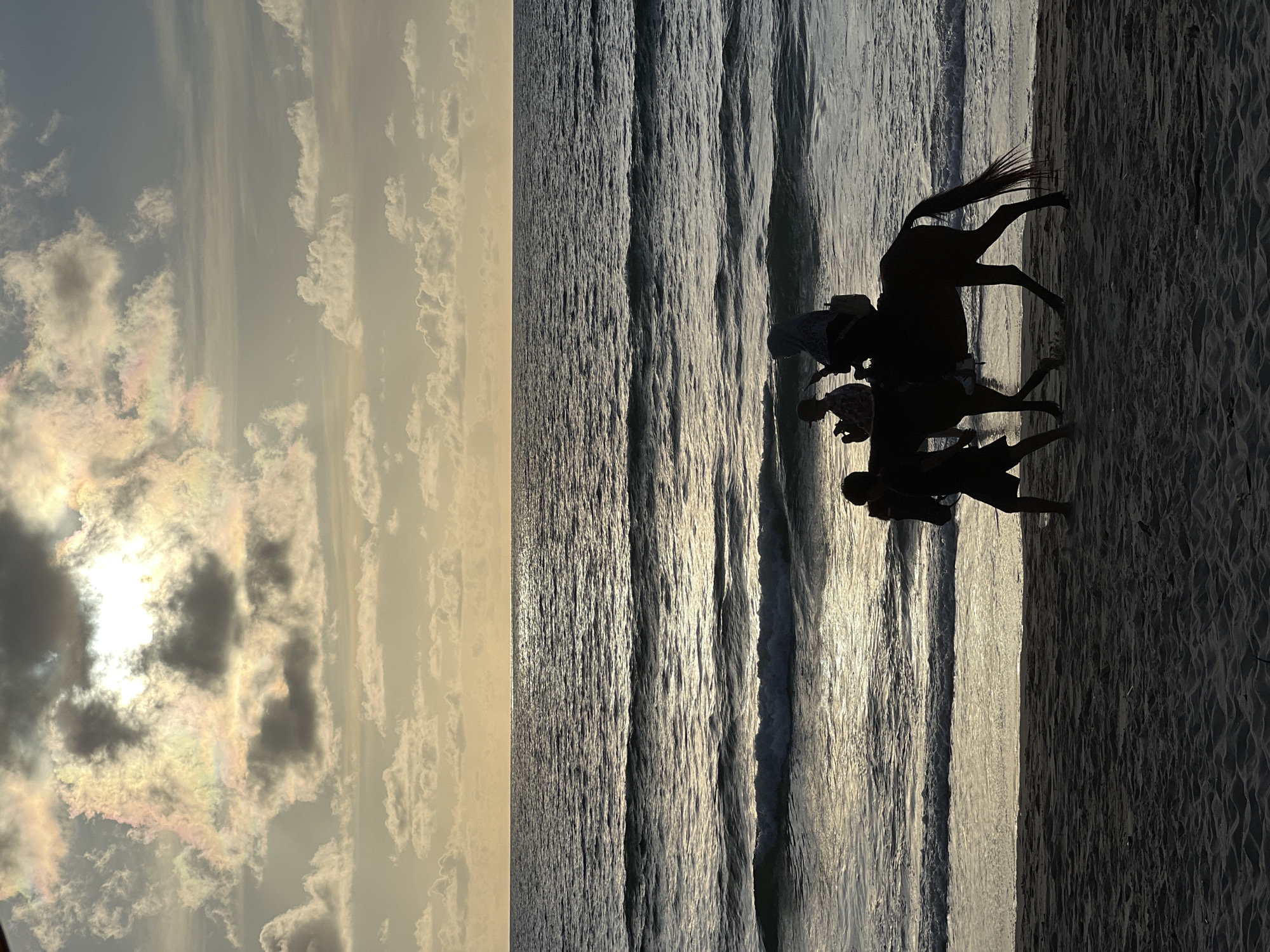 A horse is walking along the beach with three people. Two are riding the horse, and one is walking beside it. The horse and people are silhouetted against the ocean and sky. The sun is low in the sky, casting a warm glow on the clouds. The water is calm, with small waves rolling onto the shore. The sand is dark, and there are some ripples from the wind. The scene appears to be a peaceful evening on a tropical beach. The location is specified as Choeng Thale, Thailand.