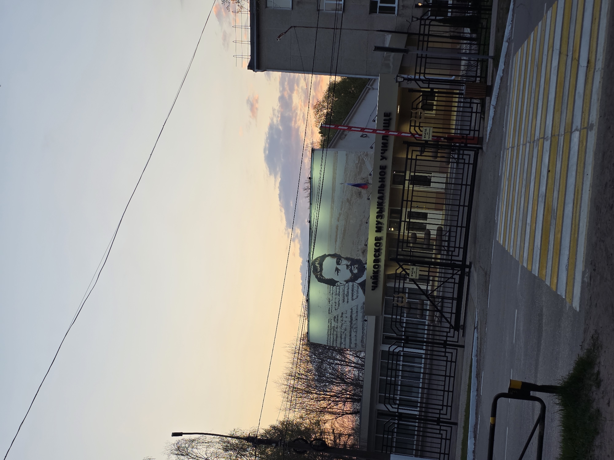 The image shows the facade of the Tchaikovsky Musical College in Chaikovsky, Russia. A large portrait of Pyotr Ilyich Tchaikovsky is visible on the building's front wall, along with Cyrillic text. The building is enclosed by a metal fence, with a crossing for people in front. A barrier is in place near the gate. Bare trees are present in front of the building. The sky transitions from a pale blue to orange.