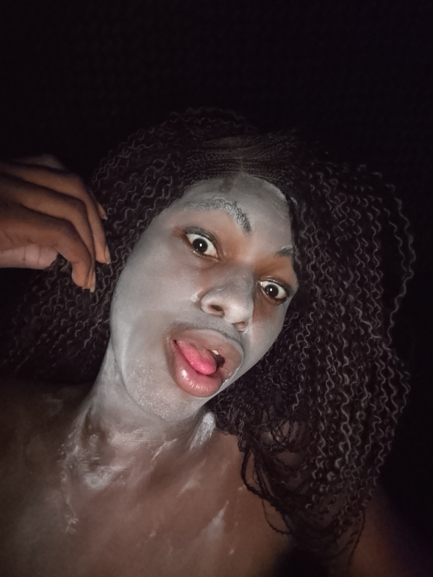 A close-up shot shows a person with dark skin and braided hair, wearing a white mask on their face and chest. They are sticking out their tongue. The background is dark. The location is Unknown, Unknown.