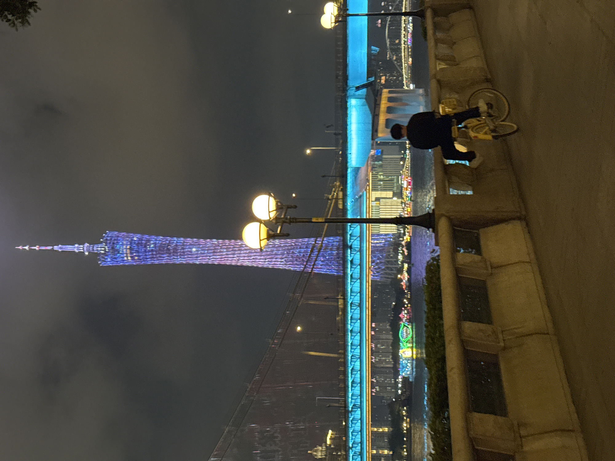 A nighttime shot shows the Canton Tower in Tianhe District, Guangzhou, China, colored with a blue and purple hue. The image is taken from a vantage point near a bridge adorned with blue lighting. Along the right side of the picture is a wall where a person stands next to a bicycle. Several street lamps are visible. Reflections of city lights shimmer on a body of water.