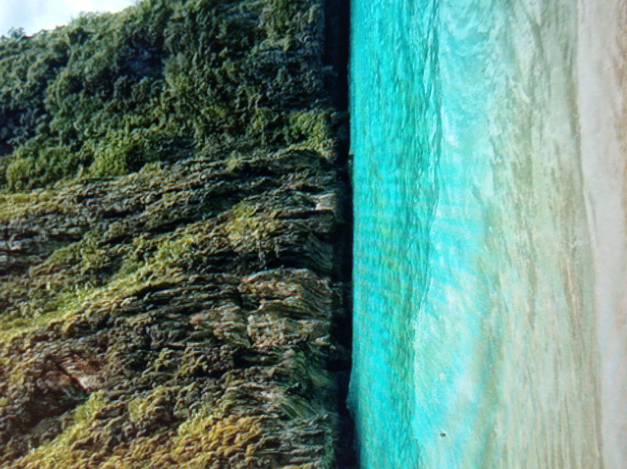 The image shows a tropical beach scene with a steep, green, and rocky cliff face on the left, bordering clear, turquoise water. The water gradually becomes lighter and more translucent towards a sandy beach, which is visible in the lower portion of the image. The environment appears to be a natural, uninhabited landscape. There are no people or man-made objects visible in the scene. The image does not depict any location in the city of Mukachevo, Ukraine.