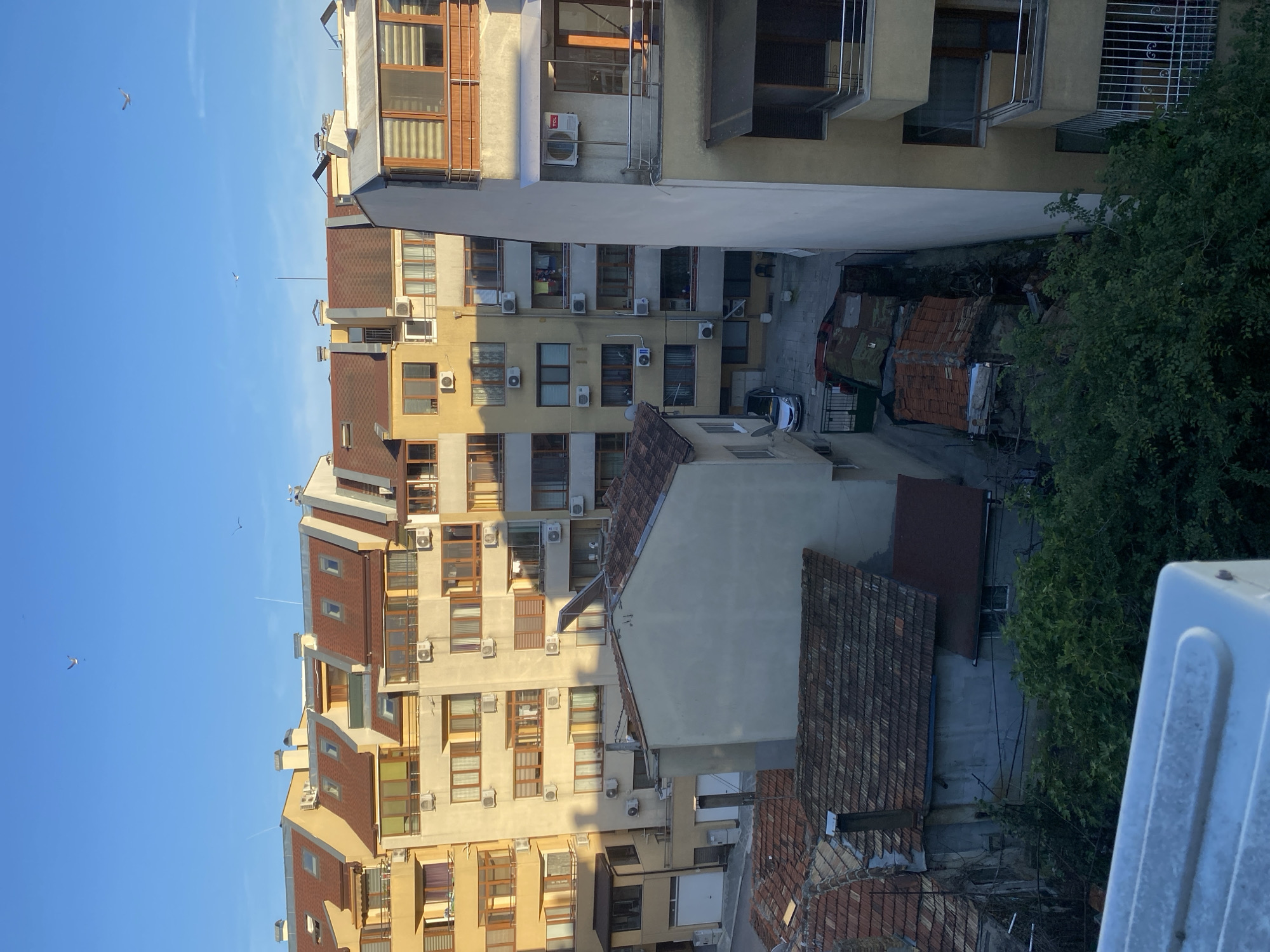 The image depicts an urban courtyard scene in Varna, Bulgaria, likely taken in the late afternoon due to the warm, angled sunlight casting shadows on the buildings. The scene is dominated by a complex of apartment buildings with a mix of architectural styles and ages. One prominent building features a beige facade with numerous windows framed by brown wooden trim. Another, taller building has a lighter facade and a distinctive reddish-brown tiled roof, with several dormer windows. Numerous air conditioning units are visible on the exterior walls of the buildings, suggesting a residential area.
In the foreground, to the right, a large, light-colored structure, possibly a rooftop unit or the side of a building, occupies a significant portion of the frame. Below this, a dense green tree adds a touch of nature to the urban environment. The courtyard itself is partially visible, revealing a paved area with what appears to be a few parked cars and small, rudimentary structures with corrugated metal and tiled roofs, suggesting storage or outbuildings. The sky is a clear, bright blue, with a couple of distant birds visible in flight, adding a sense of openness. The overall impression is one of a densely populated residential area bathed in the golden light of the setting sun. No people are visible in the frame.