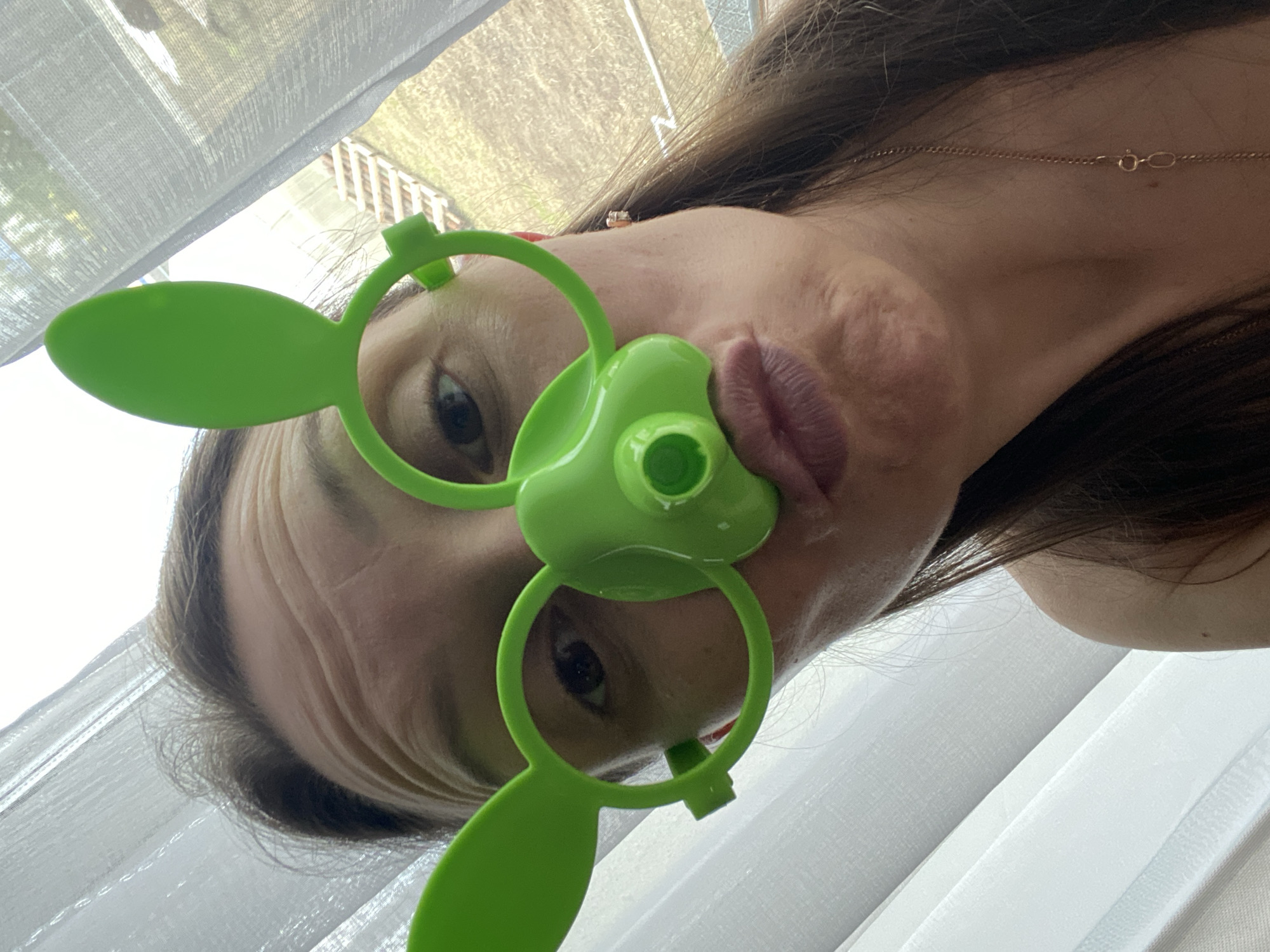 A woman is posing for a selfie. She is wearing green novelty glasses with large bunny ears on each side. The glasses cover her eyes, and the bridge of the glasses is a large, green, cylindrical object covering her nose and upper lip. The woman has long brown hair and is wearing a gold necklace. The background includes a white curtain and what appears to be foliage visible through a window. The location is unknown and cannot be determined based on the image.