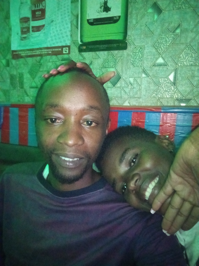 The scene depicts an indoor setting in the city of South Kamagambo ward, Kenya, characterized by a pervasive green ambient light. The background wall features a repeating geometric pattern and a lower section adorned with horizontal red and blue stripes. On the upper left wall, a poster for "DALLAS GIN" is visible, displaying two bottles. To its right, a green wall-mounted object with a dark silhouette design is present.
Two individuals with dark complexions are positioned in the foreground. The male, situated on the left, has a shaved head, a short beard, and wears a dark long-sleeved shirt with a visible lighter collar. He looks directly at the camera with a slight smile. His left hand is placed on the head of the second individual.
The second individual, partially visible on the right, leans against the male's left shoulder. This individual has short dark hair and looks towards the camera with a wide smile, revealing teeth. Their right hand, with light-colored nail polish, grips the male's left arm. The physical contact and close proximity between the individuals indicate a familiar interaction.