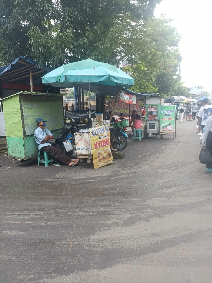 The image shows a street scene in Purwokerto, Indonesia, located at the coordinates -7.417 latitude and 109.249 longitude. A man sits on a chair in front of a food stall, waiting for customers. The stall is under a large blue umbrella and is selling "Cikur Timun Nyida", which appears to be a type of food. In the background, there are other stalls and people walking by. One of the stalls is selling "Es Pisang Hijau", which may be a type of banana-based dessert. The environment is urban, with the scene taking place on a paved road. The sky is overcast with clouds. Several people are walking down the street. There are various food stalls set up along the street with people standing around. The man sitting is likely a vendor. The image is taken from an angle that shows the street, stalls, and people walking by. The road is wet from rain.