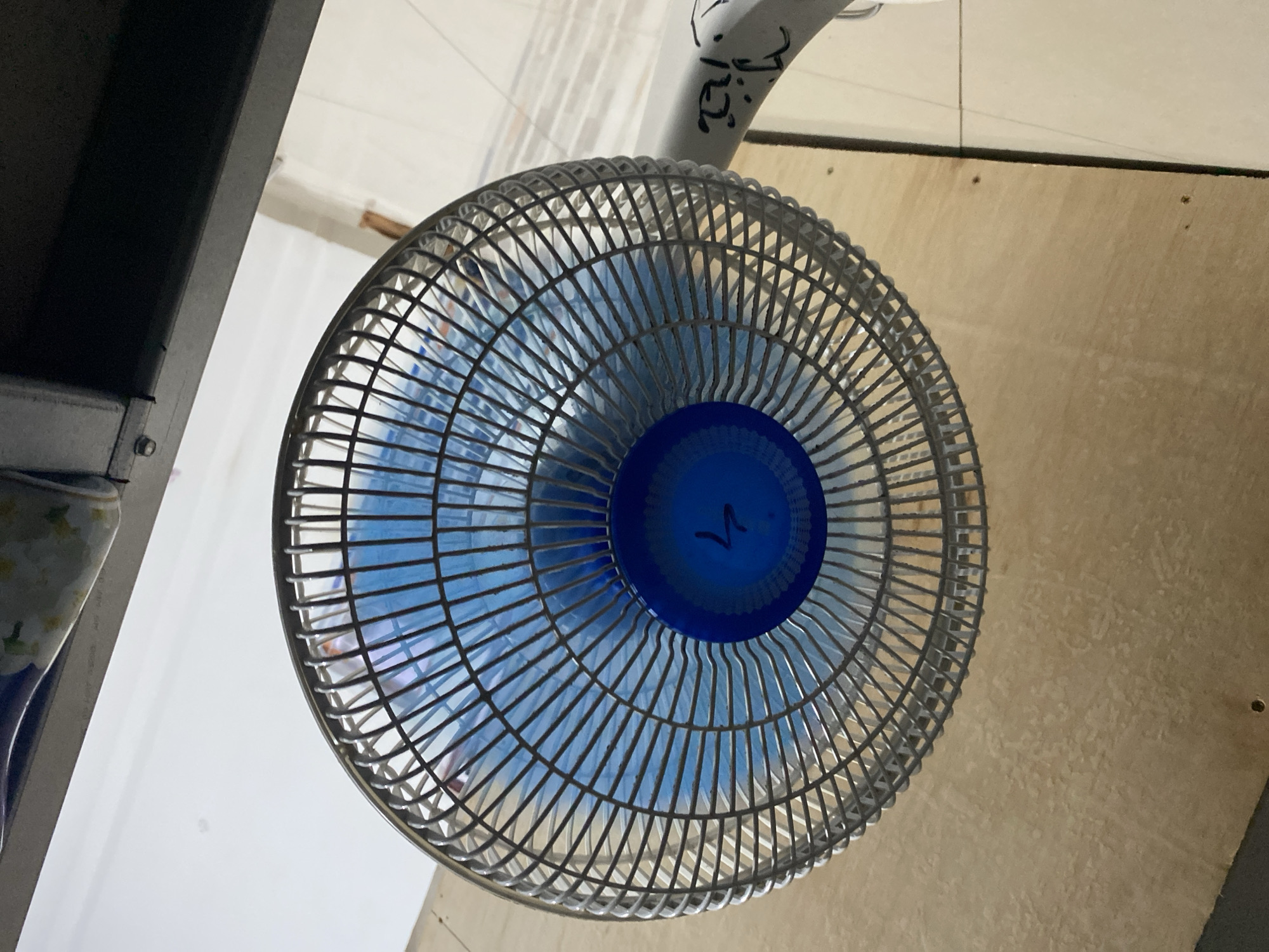A close-up, high-angle shot shows the front of an electric fan, with its metal grate and light blue blades. The fan is positioned in front of a wooden surface, possibly a wall or furniture, which appears textured and light brown. The floor below the fan is tiled with light-colored square tiles. A metallic shelf or structure is visible to the right of the fan. The overall scene suggests an indoor environment, likely a room. No specific location in Ta', Indonesia, is identifiable from the image.