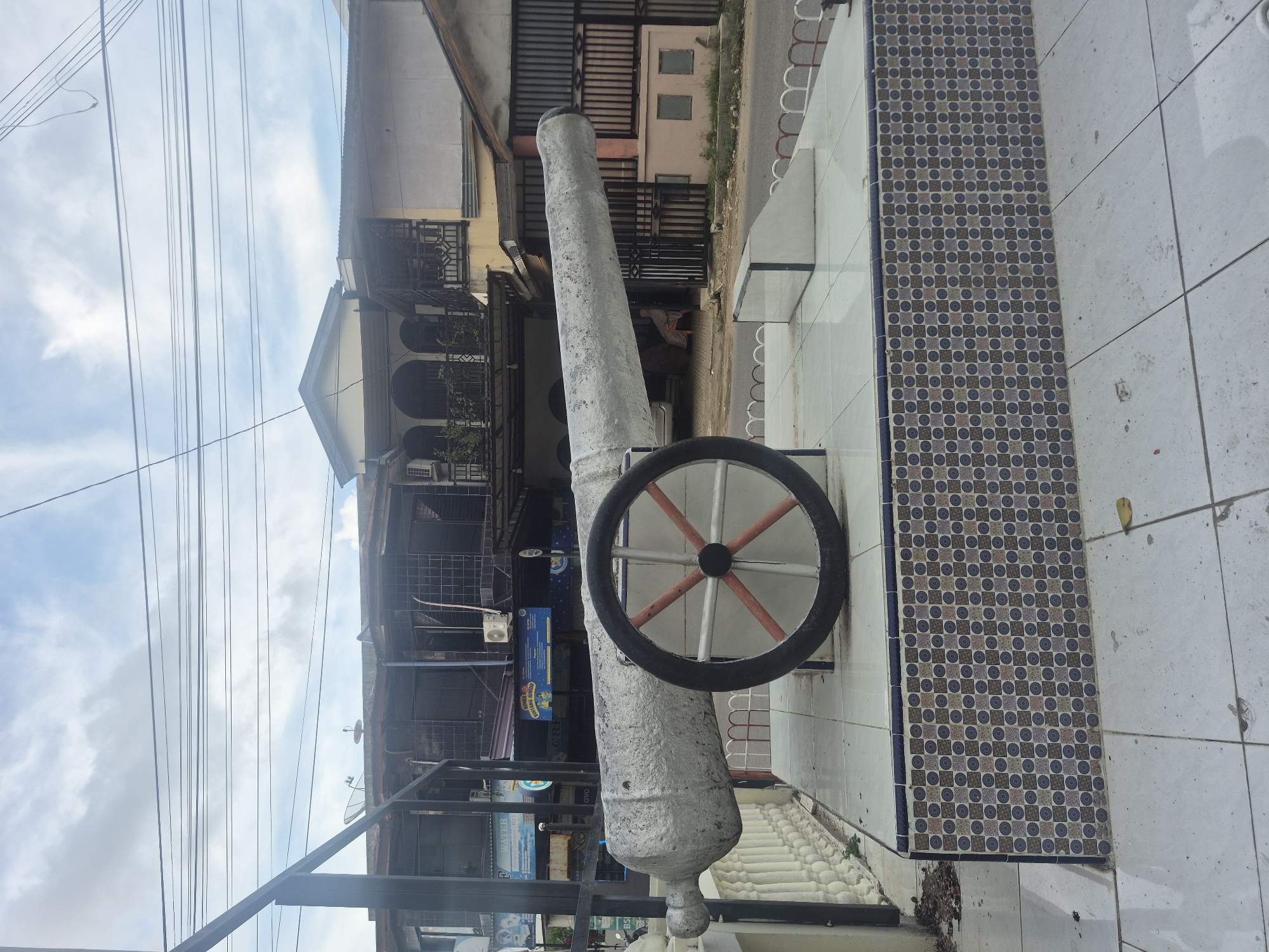 The image shows a gray cannon with a black and red wheel, seemingly a monument, positioned on a tiled surface next to a patterned tile section. The cannon faces towards buildings, including multi-story structures with balconies and shopfronts displaying signs and advertisements. Overhead, there are numerous power lines against a cloudy sky. Based on the presence of this cannon monument, likely a historical artifact, and the urban architecture, it may be positioned in the city of Lhokseumawe, Indonesia. No people are visible in the frame.