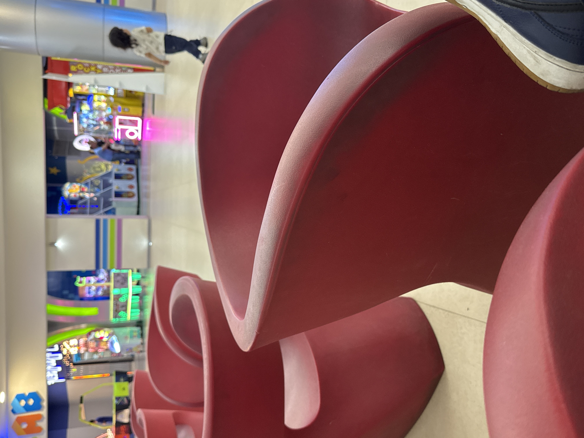 The image, taken in Yerevan, Armenia, captures an indoor public space, likely a shopping mall or entertainment complex, with a focus on modern design elements and an arcade.
In the immediate foreground, a foot wearing a blue and white sneaker with a gum sole rests casually on one of several large, abstract, red sculptural objects. These objects are deeply curved and appear to function as unconventional seating or decorative installations within the space.
In the mid-ground, a person with dark, curly hair, dressed in a patterned shirt and dark trousers, stands near a tall, silver, cylindrical architectural column. They are looking towards the background activity.
The background is dominated by a brightly lit arcade area, showcasing numerous game machines with vibrant screens and colorful lights, predominantly blues, greens, and yellows. A striking pink/magenta neon sign with stylized, possibly Cyrillic letters "ТЕ" (or "PE") glows centrally within the arcade. Towards the bottom left of the arcade, part of a sign featuring blue and orange letters, possibly "HB" or "ЯB," can be seen.
The scene is illuminated by ample artificial indoor lighting, complemented by the intense glow from the arcade games, creating a lively and engaging atmosphere typical of a leisure environment. No specific time of day is discernible beyond being indoors.