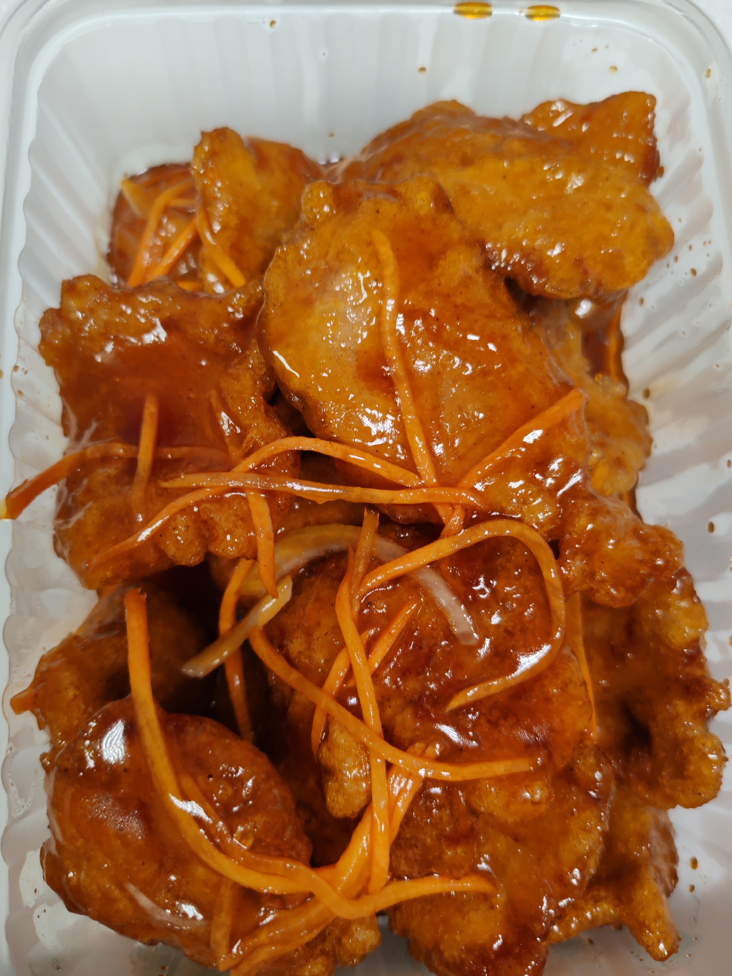The image shows a close-up, overhead view of a clear plastic container filled with pieces of battered and fried meat coated in a glossy, orange-brown sauce. Thin, bright orange julienned carrots are interspersed throughout the dish, with a few slivers of white onion also visible. The meat appears to be cut into bite-sized pieces, and the sauce has a slightly viscous quality, clinging to the food. The container is white and ribbed on the inside, a common type for takeout food. There are no people or external environmental elements visible in the image, suggesting it is a still-life shot of food. The image does not contain any information or context to place it in the city of Komsomolsk-on-Amur, Russia.