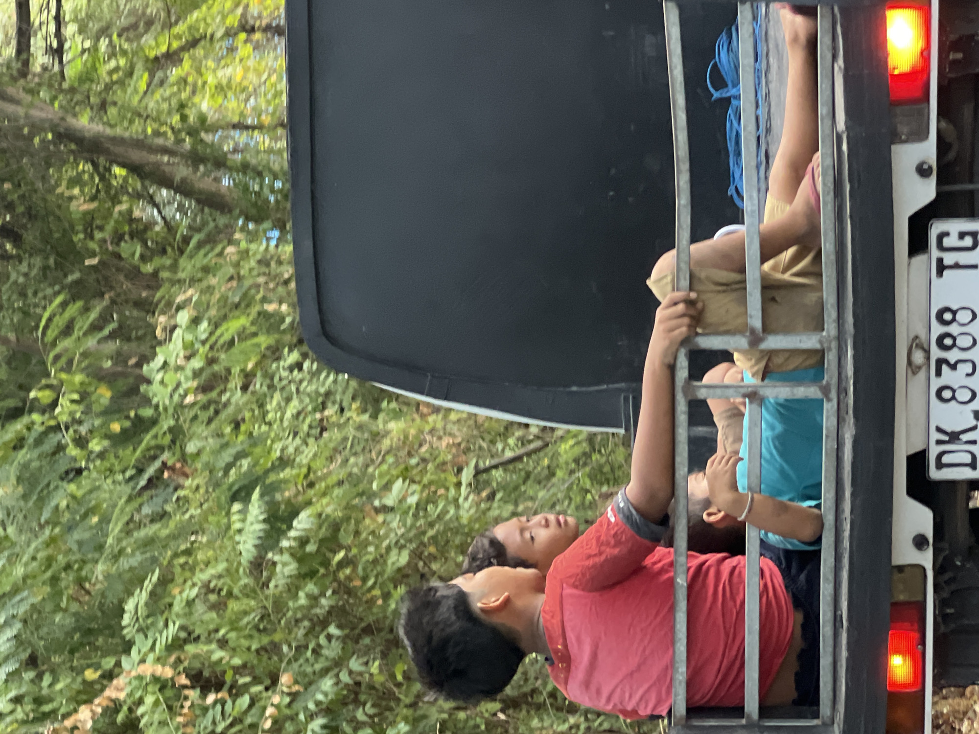 In Abang, Indonesia, two young boys are seated in the back of an open truck. The boy in the foreground, wearing a red t-shirt, is looking to his left. Behind him, another boy, wearing a blue shirt, is lying down and appears to be resting. The truck is surrounded by lush green foliage, suggesting a rural or natural setting. The truck has a license plate that reads "DK 8388 TG". The overall scene depicts a moment of travel or rest for the two children.
