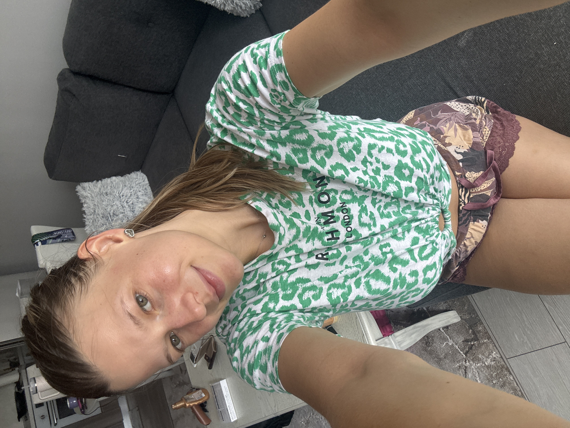A young woman with light brown hair pulled back in a ponytail is taking a selfie in what appears to be a living room. She is wearing a green and white leopard print t-shirt tied at the waist, and patterned shorts with lace trim. Behind her, a dark gray sofa is visible with a grey fluffy cushion on the left. To her left, a white table is present with various items on it, including what looks like a hairbrush and some papers. The floor is a mix of wood and carpet. The image does not provide any specific indicators that would suggest a location in Sofia, Bulgaria.