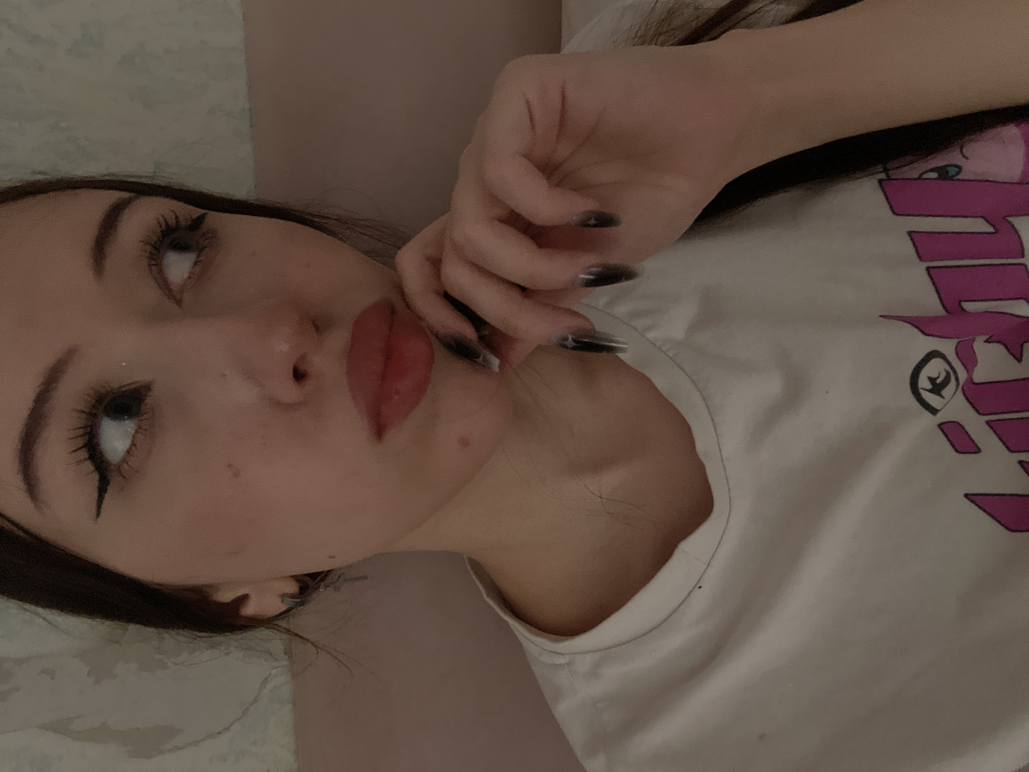 The image shows a close-up of a young woman's face and upper body. She has dark hair, winged eyeliner, and is wearing a white t-shirt with a graphic print. Her right hand is touching her chin. The background appears to be a textured wall. The location of this image in Votkinsk, Russia is unknown and cannot be determined from the visual information provided.