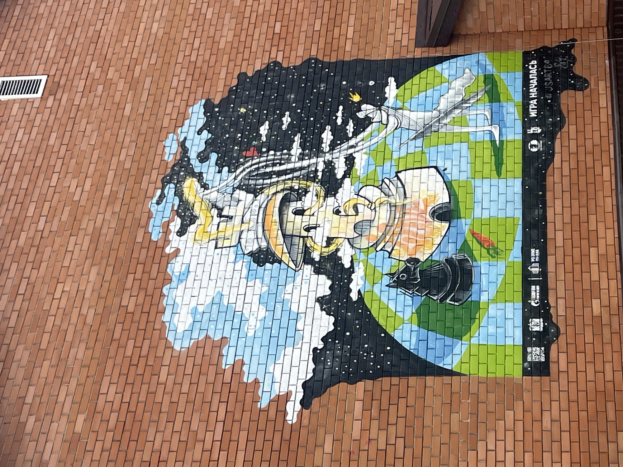 This media file is a photograph of a mural painted on a brick wall in Mezhdurechenskiy, Russia. The mural is a colorful and abstract depiction of space and Earth. The left side of the mural shows a swirling galaxy with stars and nebulae, rendered in black, white, and blue. Emerging from this cosmic scene is a stylized, metallic object that resembles a complex piece of machinery or a spacecraft, with gears and pipes visible. Yellow and orange hues highlight parts of this object.
The right side of the mural transitions to a checkered pattern of green and blue, representing the Earth or a similar planet. Across this celestial landscape, a white, flowing form resembling a ribbon or a wing stretches from left to right. A silhouette of a bird or an angel with outstretched wings is subtly integrated into this form. There is also a small, orange carrot depicted on the green checkered section. A black object that looks like a vintage camera or projector is positioned in the lower right quadrant of the Earth section.
The mural is painted on a red brick wall. In the upper left corner of the photograph, a white ventilation grate is visible on the brick wall. On the far right edge of the mural, there is a vertical black strip containing white text and symbols. The text reads "ИГРА НАЧАЛАСЬ" which translates to "The Game Has Begun" in Russian. Below this are several small icons and the hashtag "#USARTCO". A QR code is also present at the bottom of this black strip.
The overall impression of the mural is one of transition and exploration, with elements of the abstract and the tangible. The lighting suggests it is daytime, with diffused light, but no specific time of day or weather conditions are evident. The mural appears to be located outdoors, on the side of a building.