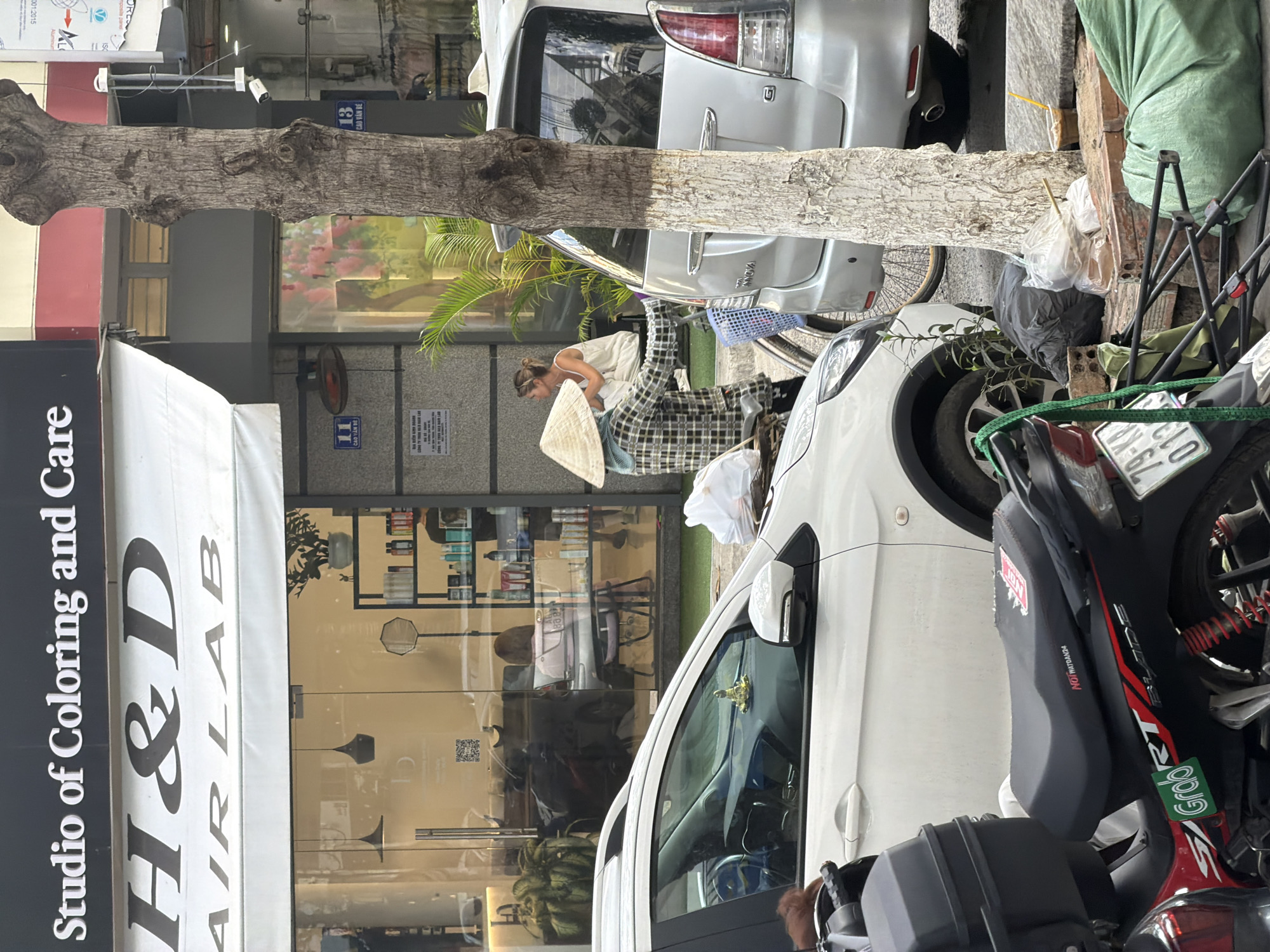 The image depicts a street scene in Bac Nha Trang Ward, Vietnam, characterized by a row of businesses and parked vehicles. In the foreground, a red and black motorcycle with a "Grab" logo is parked. To its right, the rear of a silver SUV is visible, and to the left, a white car is parked. A young woman wearing a conical hat and a checkered shirt is seen next to a bicycle, appearing to be interacting with another woman in a white dress who is sitting on a chair inside the salon. The salon, identified as "H&D HAIR LAB" and "Studio of Coloring and Care," has a glass facade revealing shelves stocked with beauty products. A tree trunk with white paint marks stands prominently in the mid-ground. The ground is paved, with some debris and plastic bags scattered near the tree. The overall impression is one of a typical urban street with commercial activity and pedestrian interaction.