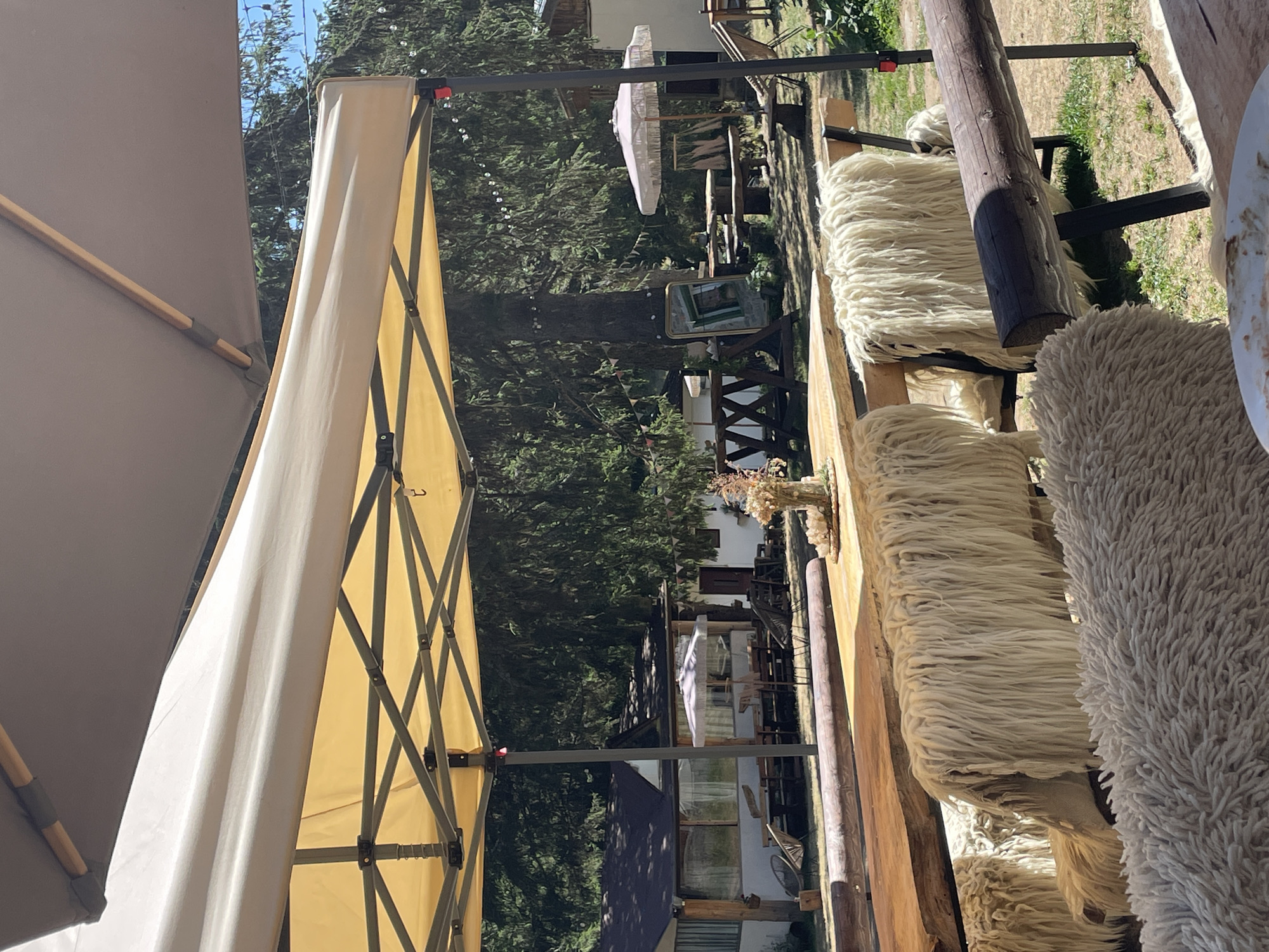 The image depicts an outdoor seating area at a restaurant or cafe, likely located in or near Smolyan, Bulgaria, suggested by the rural setting and traditional elements like sheepskin throws. The scene is set under a large, yellow tent structure, with another darker tent partially visible overhead. In the foreground, a long, rustic wooden table is set with a white plate and a floral centerpiece. Several wooden benches are positioned along the table, each draped with thick, shaggy sheepskin throws, creating a cozy and rustic aesthetic.
Behind the seating area, the environment transitions into a more natural landscape. Pine trees with dense foliage dominate the background, indicating a mountainous or forested region. Several small, rustic buildings with dark roofs and white walls are visible among the trees. Some of these buildings appear to be part of the establishment, with outdoor seating areas and umbrellas. In the mid-ground, there are picnic tables and benches, and a large mirror leans against a tree. String lights are visible across the trees. The ground is covered with dry grass and dirt. The overall impression is one of a relaxed, outdoor dining experience in a natural, possibly rural, setting. There are no people clearly visible in the frame.
