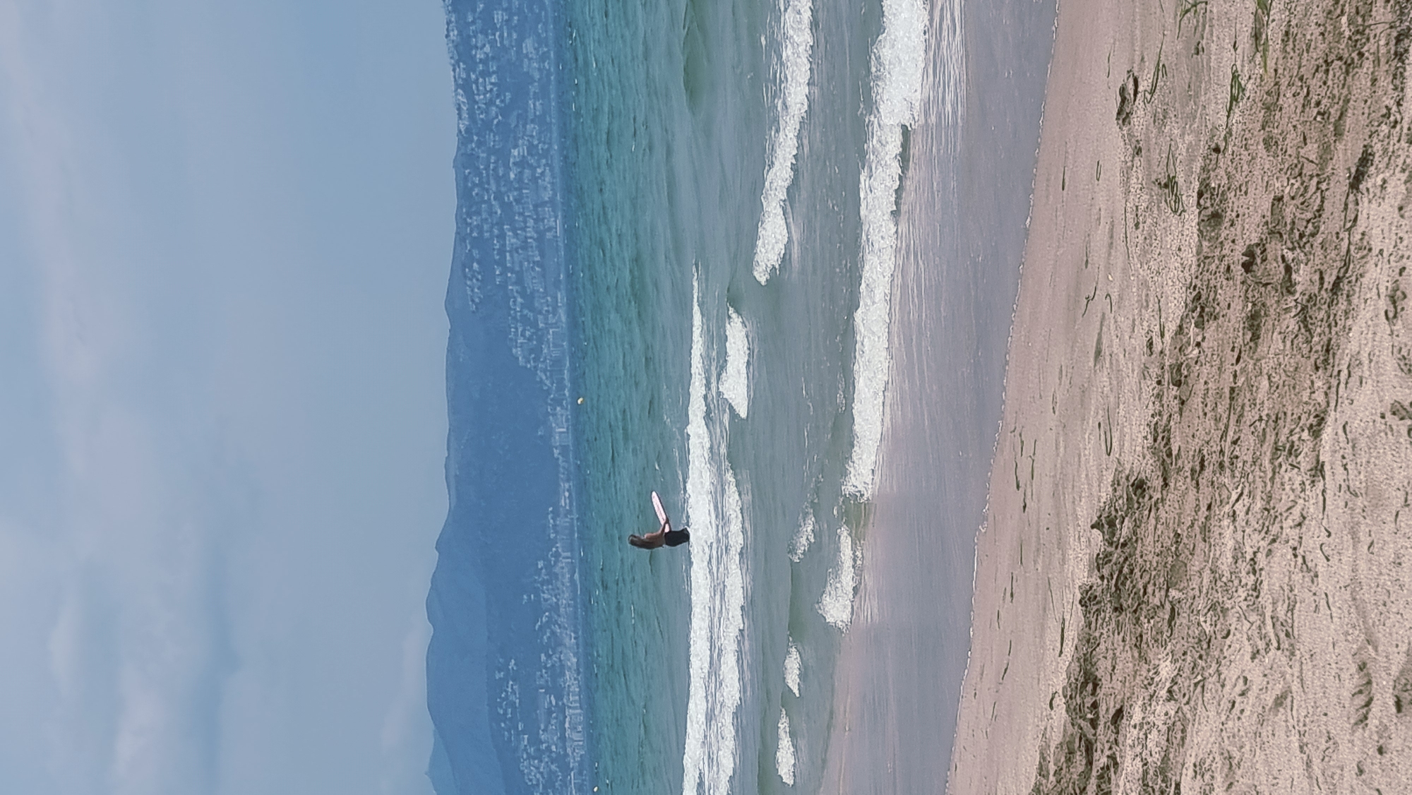 In the city of Castelló d'Empúries, Spain, on a sandy beach, a lone person stands in the shallow ocean waters, holding a pink and white surfboard. The person is facing away from the camera. The water is a turquoise blue with white-capped waves rolling towards the shore. In the distance, a range of blue mountains with a town built along their slopes are visible under a cloudy sky. The foreground is dominated by the sandy beach, with some dry seaweed scattered on the sand. The scene depicts a leisurely day at the beach with water activities.