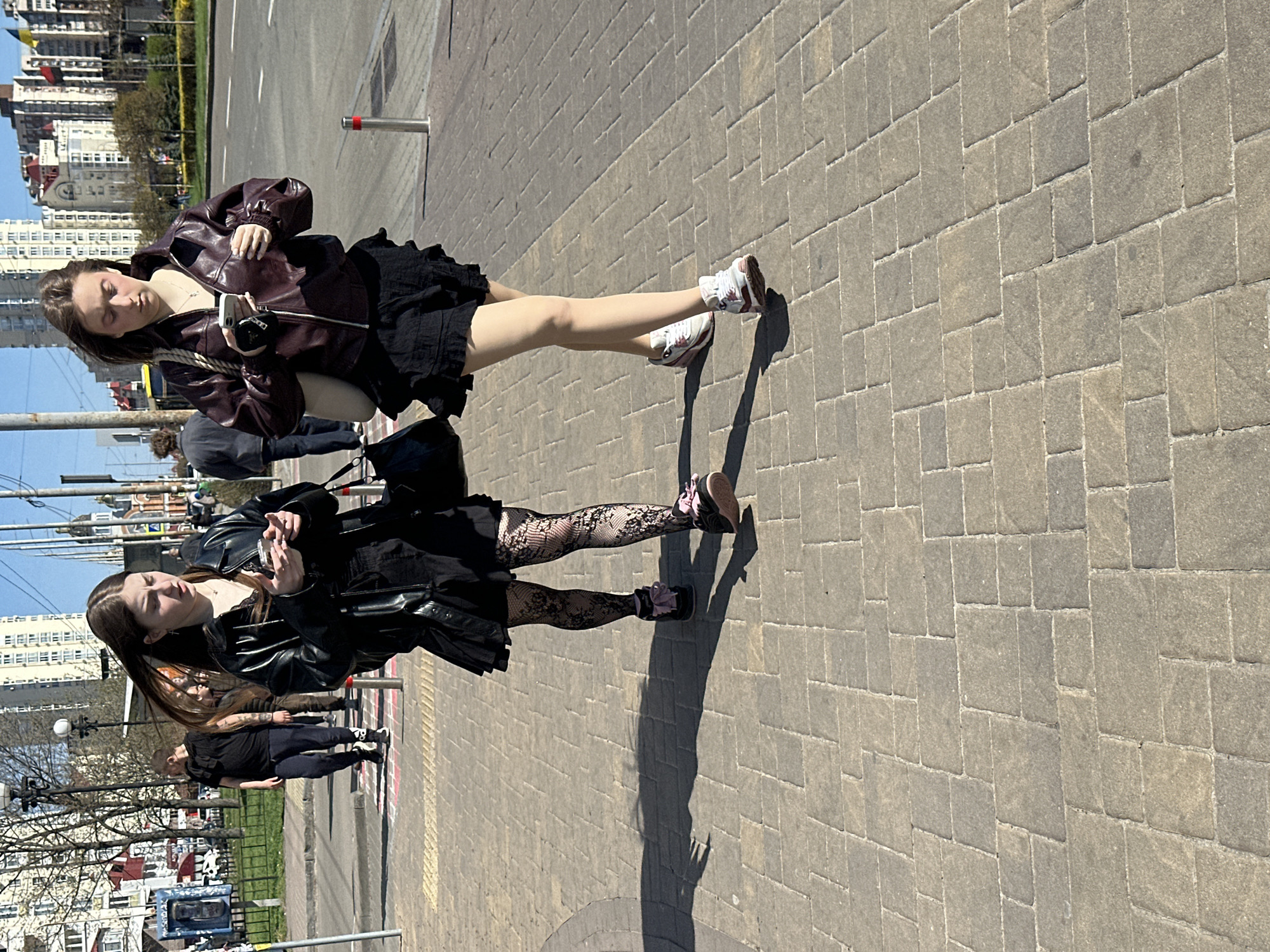 Two young women stand on a paved sidewalk in Kyiv, Ukraine. The woman on the left wears a black dress, black leather jacket, and patterned tights, holding a cell phone. The woman on the right wears a black skirt, maroon leather jacket, and white sneakers, also holding a cell phone. Buildings are visible in the background, along with trees, streetlights, and a road. Other pedestrians are present in the background. The weather appears sunny.
