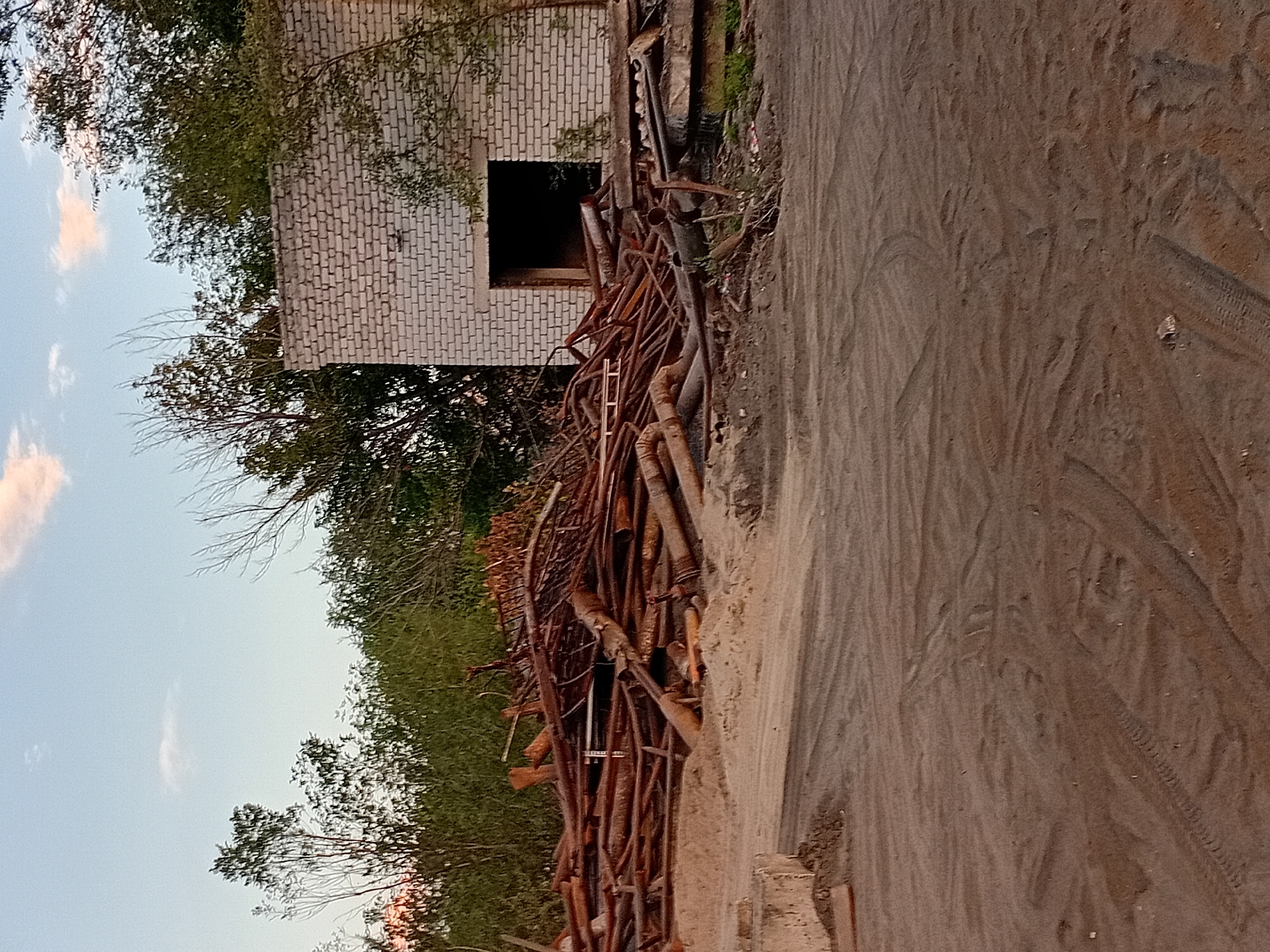The image depicts an outdoor scene with a pile of rusty pipes and debris in the foreground. Behind the debris is a partially constructed or dilapidated building made of white brick, with a dark, open doorway. Trees and vegetation are visible behind the building and to the left. The ground is covered in sand and dirt, with tire tracks visible, suggesting recent vehicular activity. The sky is partly cloudy, with hints of sunset colors. The scene appears to be in an industrial or construction area. This location is believed to be in the city of Semey, Kazakhstan.