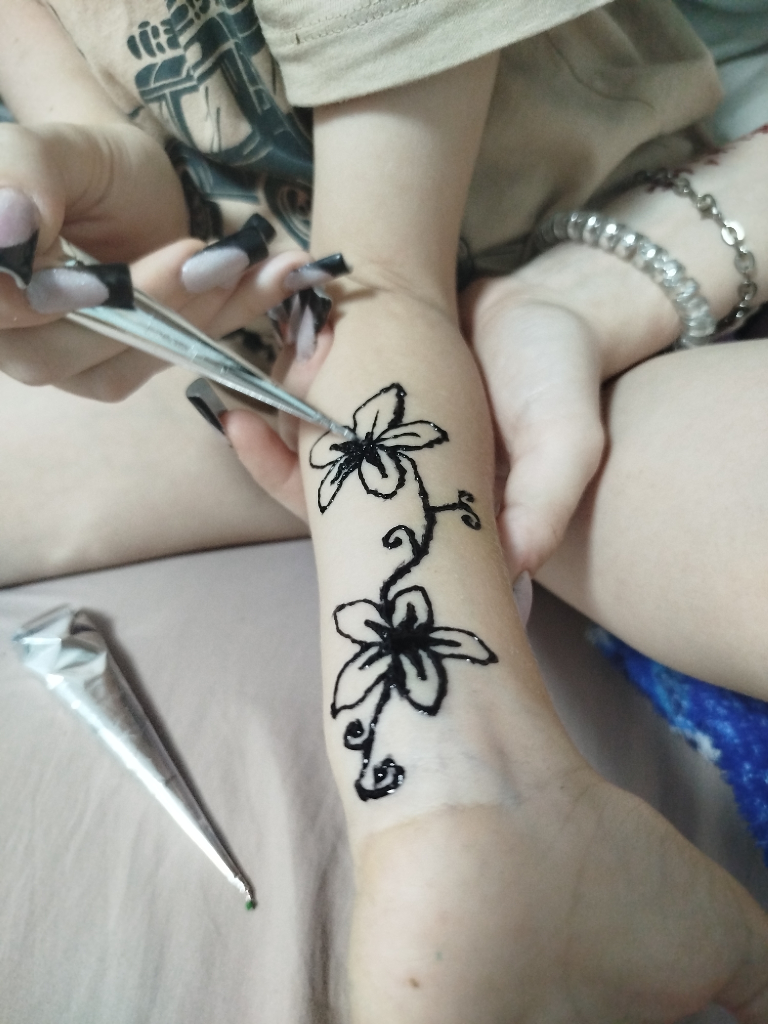 In a close-up, indoor shot, a person is applying black henna paste to their forearm in the form of two stylized flowers connected by a vine. The henna cone is visible in the lower left corner, with a small amount of green paste at its tip. The forearm is positioned on a beige fabric surface, possibly a blanket or bedsheet. The person's left hand, with long, dark-tipped acrylic nails, holds a pair of tweezers, though their purpose in the henna application is unclear from the image. A silver bracelet is visible on the person's right wrist. The background is blurred but suggests a domestic setting. There is no discernible information in the image to suggest a location in Zavodoukovsk, Russia.