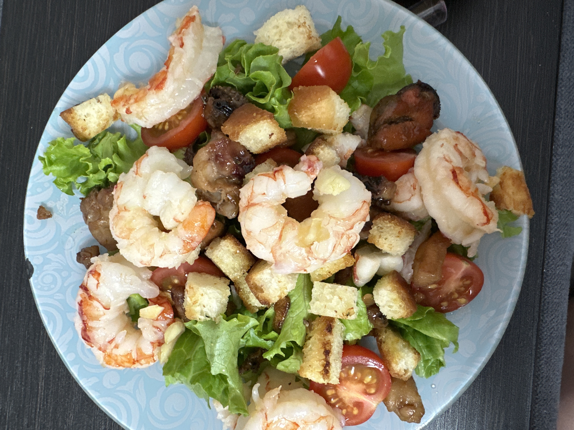 The image shows a close-up, overhead view of a shrimp salad served on a blue patterned plate. The salad contains several large, cooked shrimp, cherry tomato halves, croutons, and leafy green lettuce. Some pieces of dark, possibly meat, are also visible. The plate is set on a dark, textured surface, likely a table. The scene appears to be indoors, with no discernible city or environmental context from Chaikovsky, Russia, or any other location. The focus is entirely on the food.