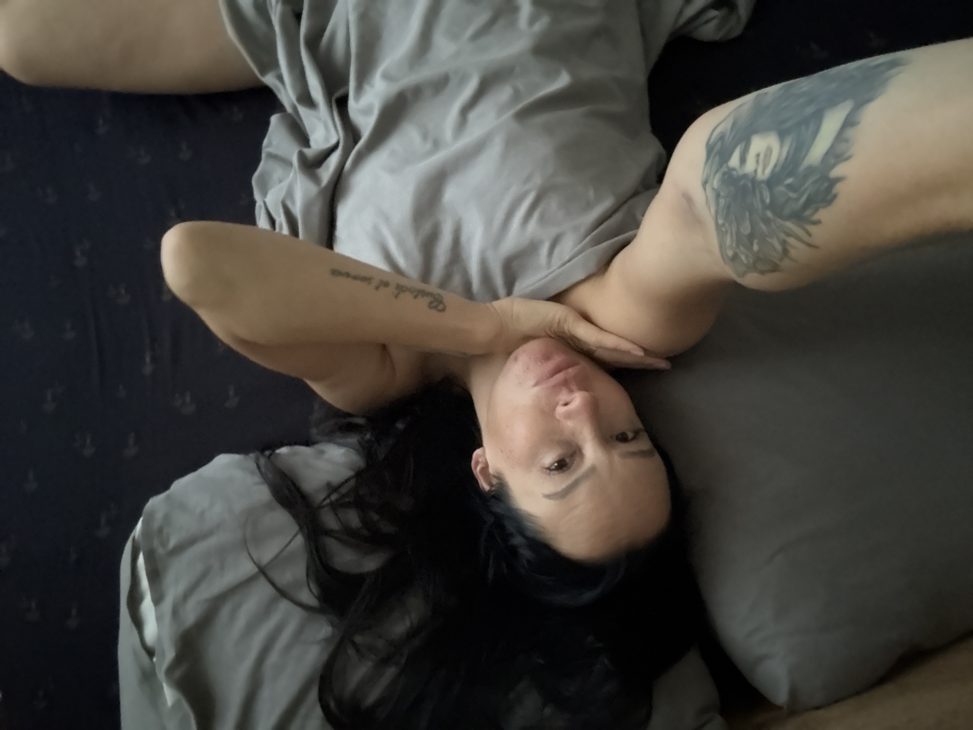 A woman with long dark hair lies in bed, looking at the camera. She is covered by a gray blanket. On her left arm, there is a tattoo of a face with wings. On her right forearm, there is a tattoo of text. The background is a bed with gray and dark blue patterned sheets. The scene appears to be in a bedroom. There is no information in the image to determine the specific location within the city of Chaikovsky, Russia.