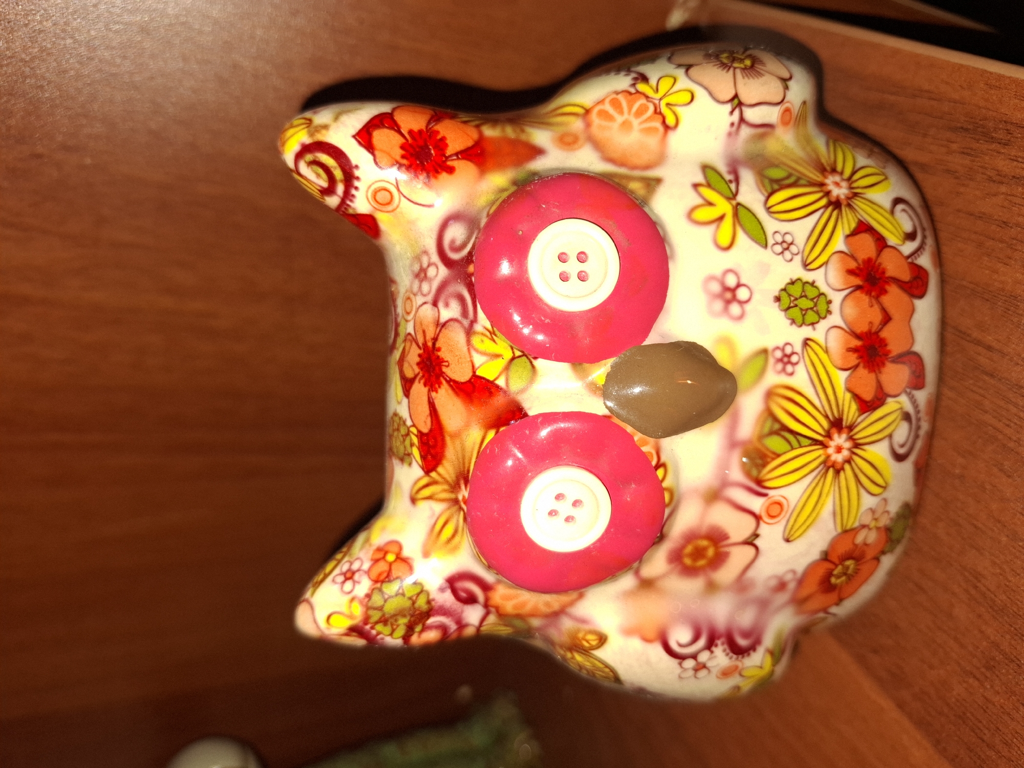 A close-up, eye-level shot shows a ceramic owl figurine with a colorful floral pattern. The owl has two large, bright pink circular eyes with white centers that have four small holes each, resembling buttons. In the center of its face is a small, glossy, brown, oval-shaped nose. The owl's body is covered in a vibrant pattern of red, orange, yellow, and pink flowers, as well as green leaves and red swirling designs. The figurine is positioned on a wooden shelf, likely in a home or store. The background is a dark wooden surface. The image was taken in Almaty, Kazakhstan.
