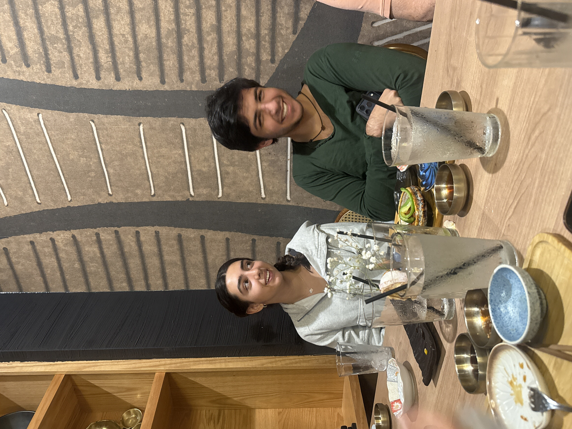 Two individuals are seated at a wooden table in what appears to be a restaurant setting. The male subject is wearing a dark green long-sleeved shirt and is smiling towards the camera. The female subject is wearing a light gray hooded sweatshirt and also smiling towards the camera.
On the table are several glasses filled with liquid and straws, as well as small bowls and plates containing food. The overall setting suggests a dining environment.
In the background is a wall with a textured pattern consisting of brown and black stripes. Wooden shelves can be seen in the bottom left corner of the frame.
The image was taken at a location in Southlake, United States of America.
