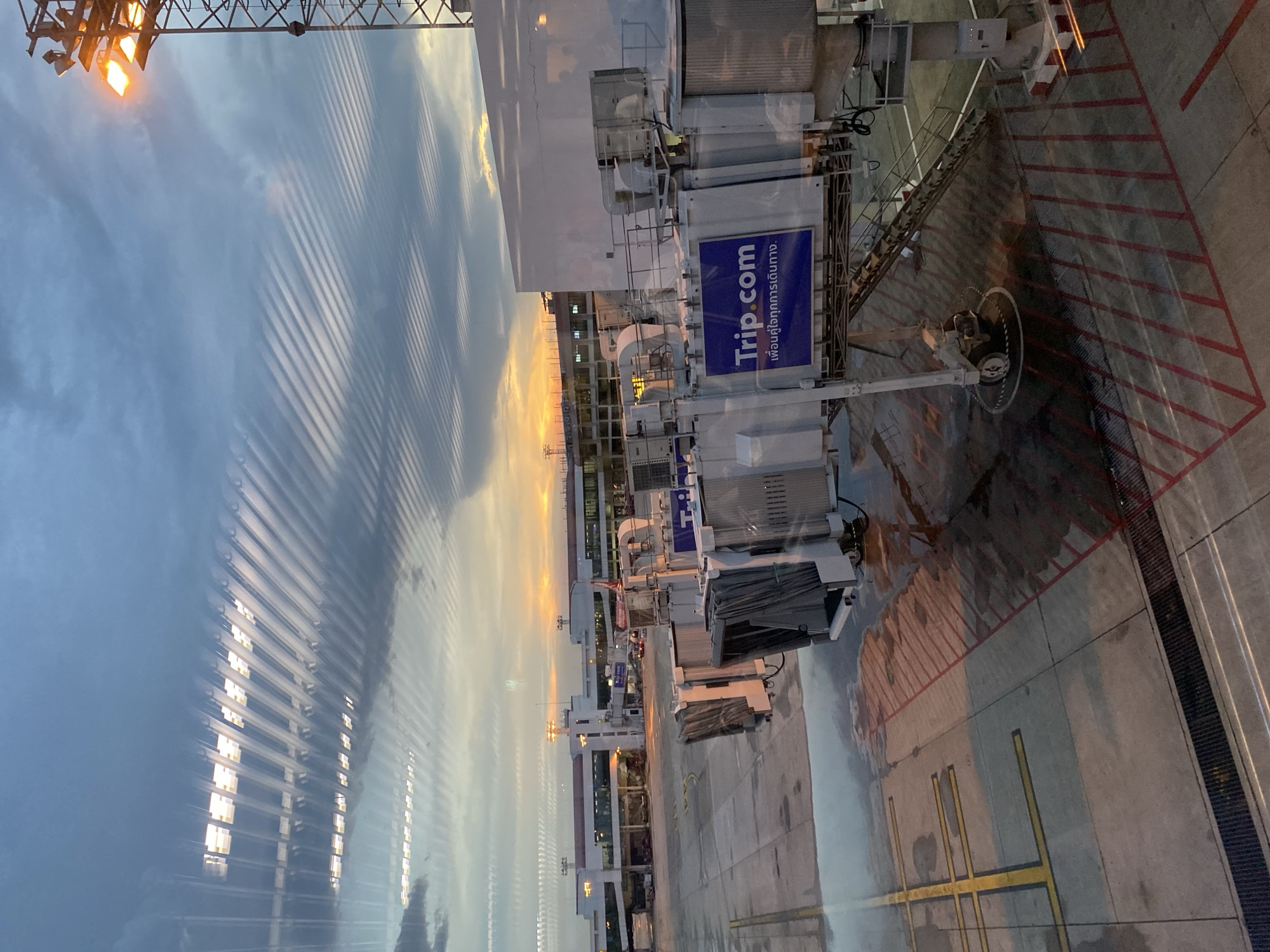 The image captures an airport scene, likely taken from inside an airplane. The scene includes an airport jet bridge extending toward a building, which hosts "Trip.com" signage, written in both English and Thai, indicative of the location. The background features an orange-tinged sky suggesting either sunrise or sunset. The foreground, likely a window, has vertical strips reflecting the scene outside. The area below the jet bridge appears to be a concrete runway with red and yellow painted markings. The image appears to be taken in Bangkok, Thailand, as the native language present is Thai.