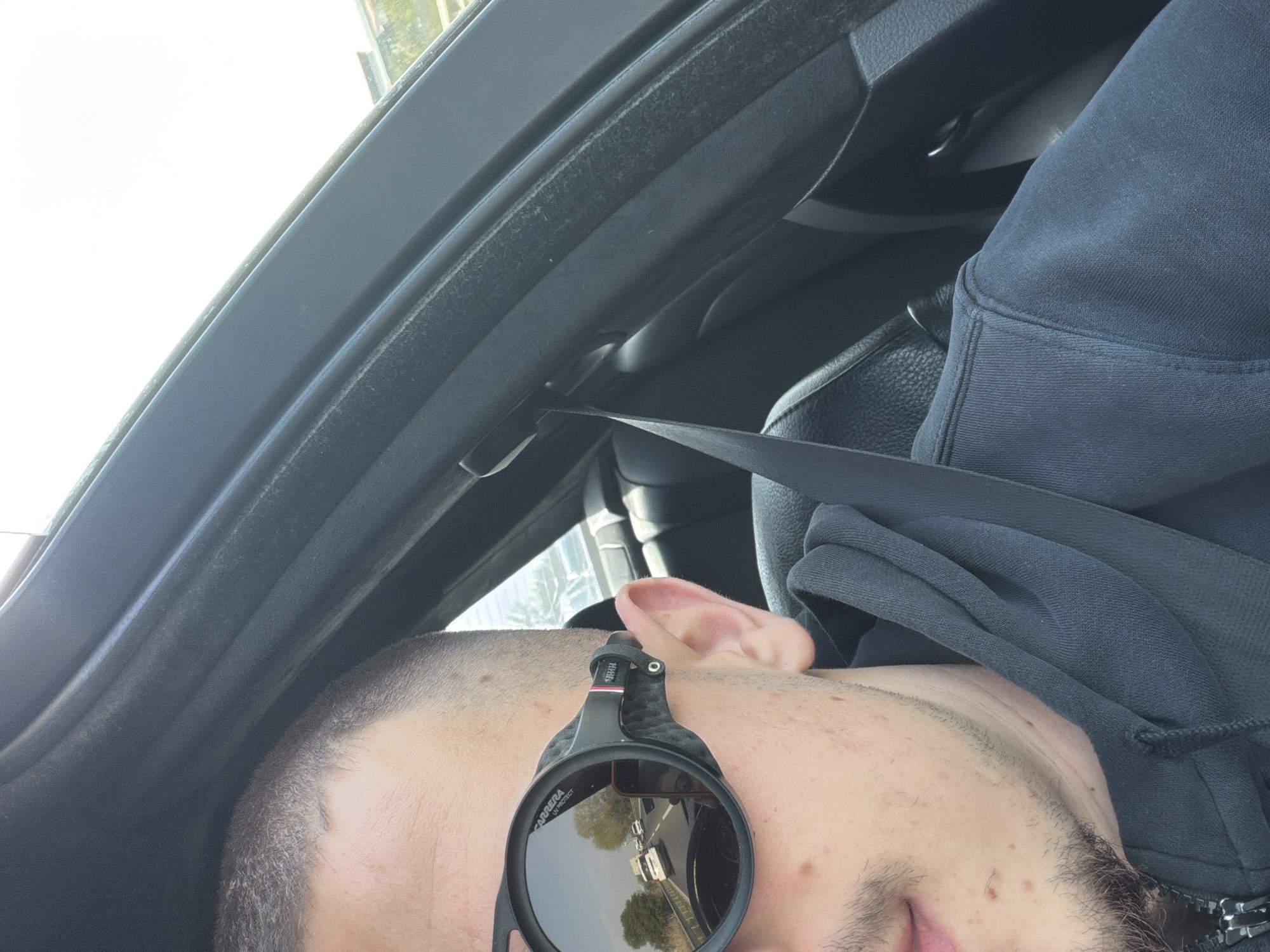 A close-up, tilted, indoor shot shows a person in a car. The person has short hair, fair skin, and is wearing sunglasses. A seatbelt crosses the person's body. The car's interior is visible, including the roof, seat, and dashboard. The sunglasses reflect a scene with trees and possibly other vehicles, but the location is not identifiable as Plovdiv, Bulgaria from the details provided.