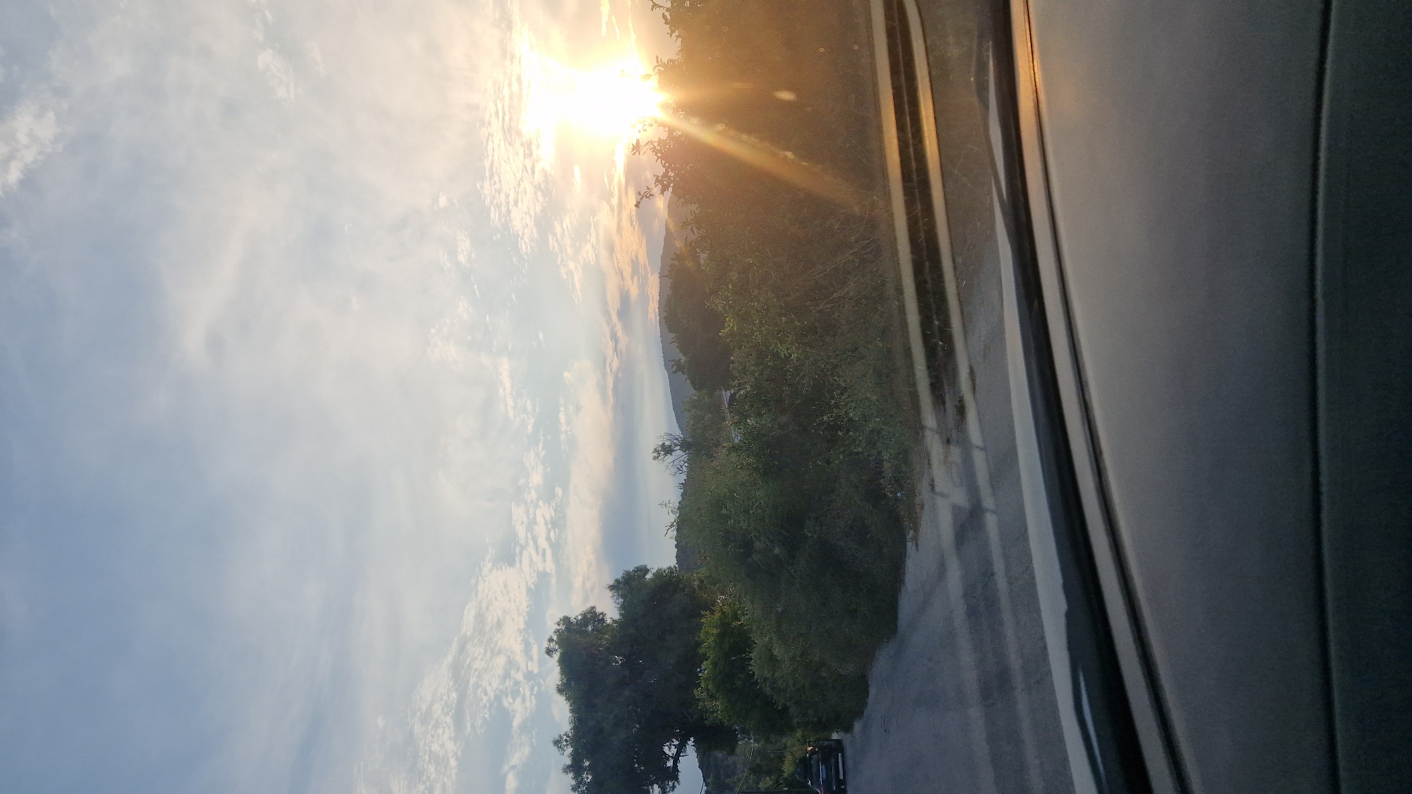 The image depicts a scenic view from the perspective of someone inside a vehicle, likely looking out through the windshield. The scene is dominated by a bright sunset, casting a warm glow over the landscape. In the foreground, the dark dashboard of the vehicle is visible, with some reflections from the road and sky.
The road, paved with asphalt, curves to the left, leading towards a dense area of green foliage and trees. A dark-colored SUV is parked on the left side of the road, facing away from the viewer. Beyond the road and foliage, a gentle slope of hills is visible, covered in greenery. The sky above is a pale blue, scattered with wispy white clouds. The sun, positioned high and to the right of the frame, is a brilliant, almost blinding source of light, with visible sunbeams fanning out across the scene.
This picturesque setting is located in the Municipal Unit of Asini, Greece. The composition suggests a rural or semi-rural area, characterized by its natural beauty and the peaceful ambiance of the sunset.