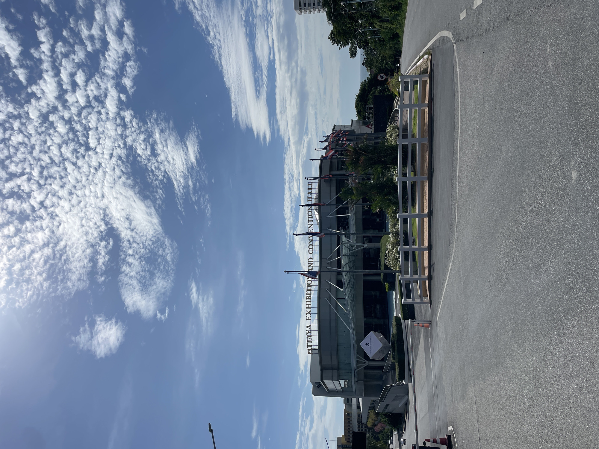 The image shows the Pattaya Exhibition and Convention Hall in Pattaya City, Thailand. The building is a large, modern structure with a glass facade and a prominent sign displaying its name. In front of the building, there are several flags flying, and landscaping with bushes and flowers. The scene is set against a bright blue sky with scattered white clouds. In the foreground, there is a road with a white fence and some traffic cones. The road curves to the right, leading away from the convention hall. The overall impression is a sunny day in a well-maintained urban environment.